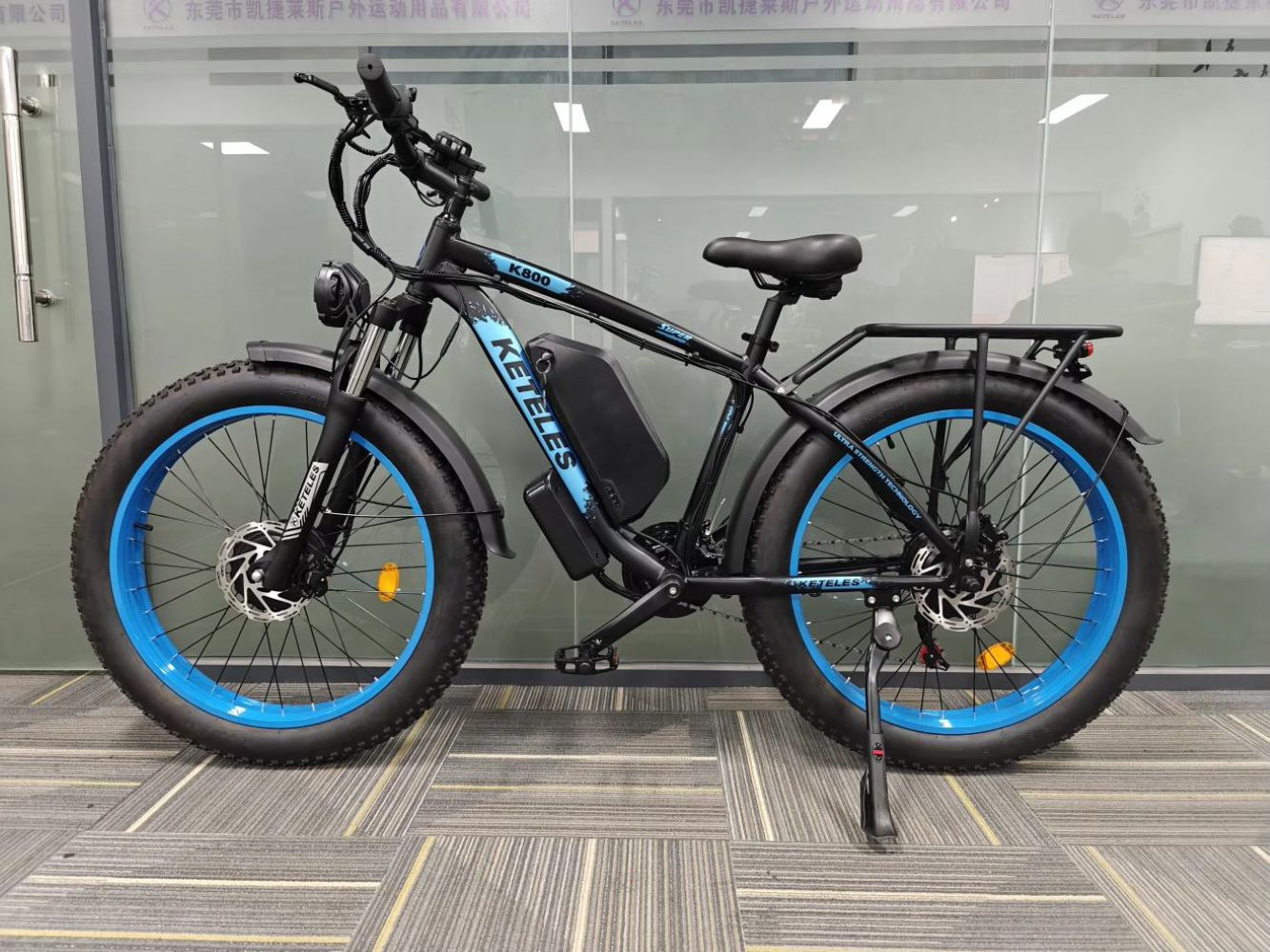 Bicicletta elettrica nera e blu. Ha pneumatici larghi, una batteria e un portapacchi. È parcheggiata su un tappeto grigio a fantasia.