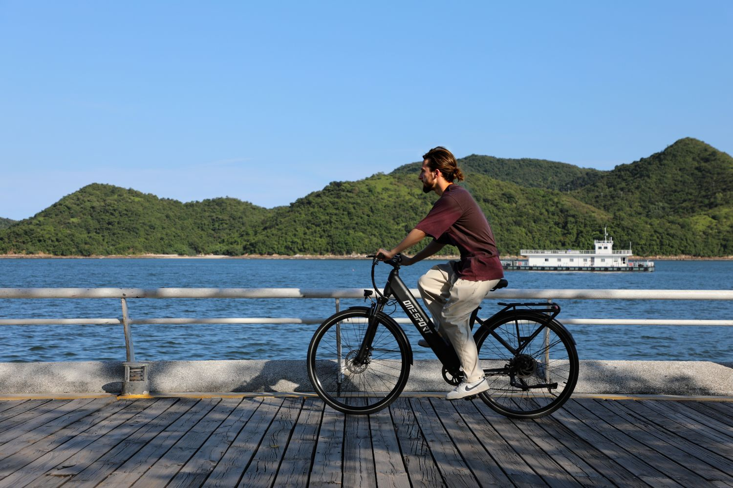 Hombre en bicicleta pedalea por un muelle de madera. Montañas y mar al fondo, cielo azul claro.