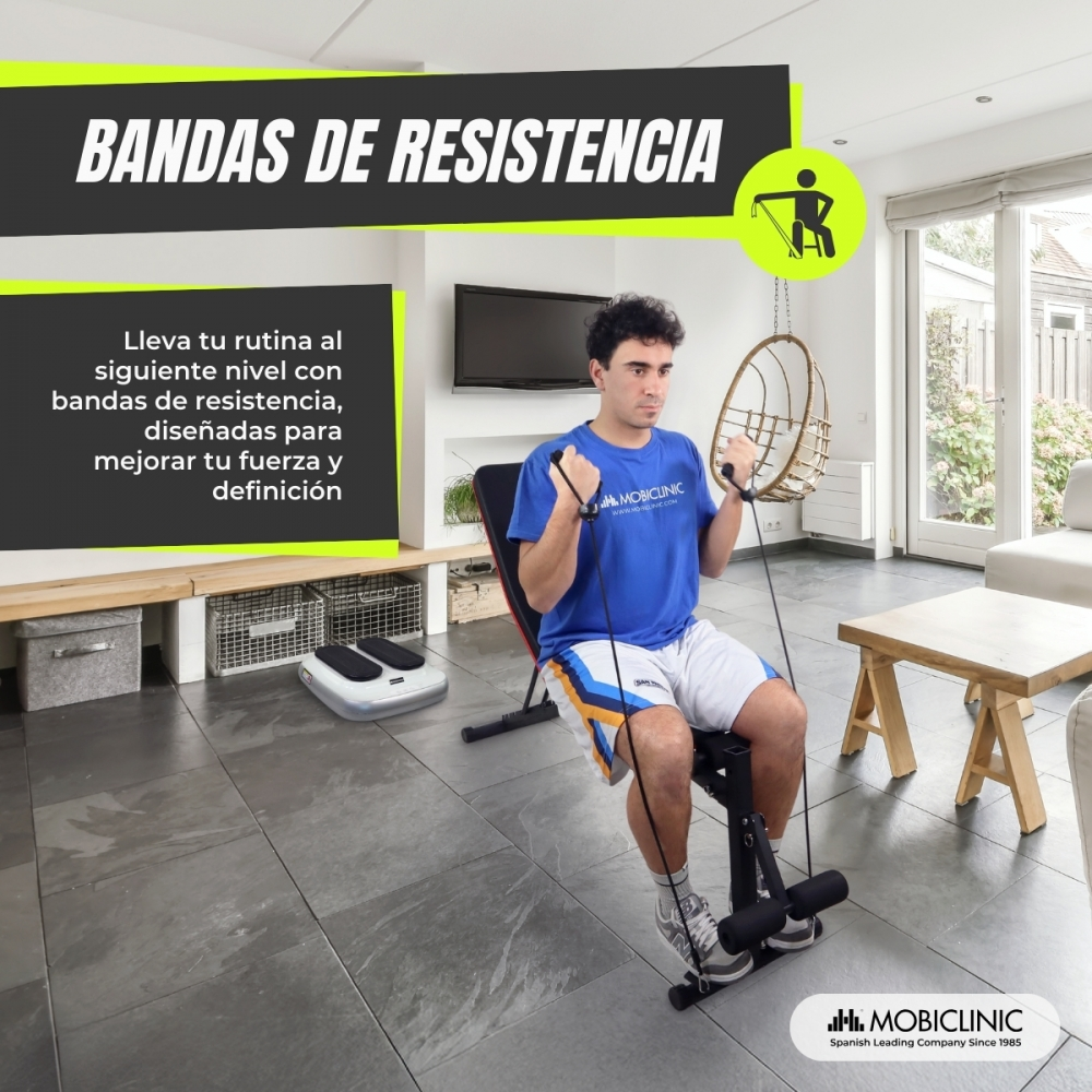 Hombre ejercitándose con bandas de resistencia en una sala de estar. Está sentado en un banco, sosteniendo las bandas. El fondo incluye muebles y ventanas.