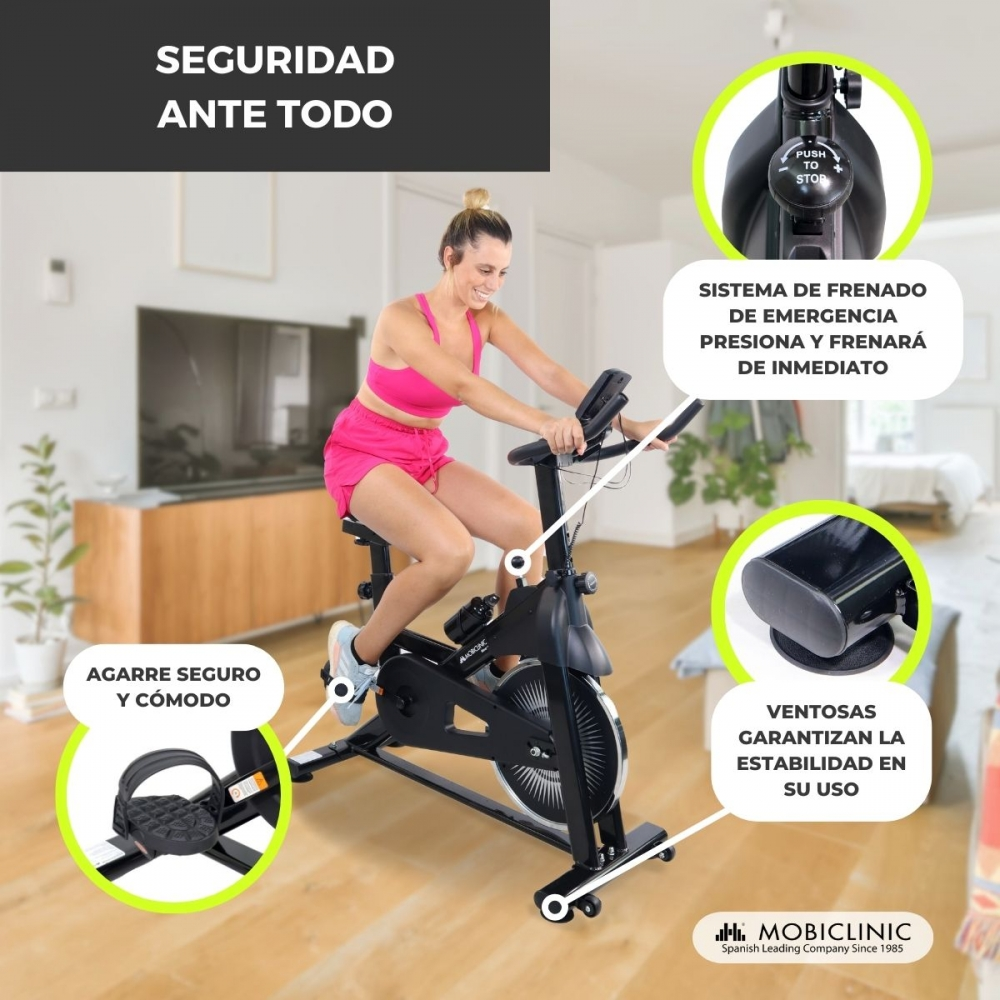 Una mujer en ropa deportiva rosa hace ejercicio en una bicicleta estática negra. Los detalles muestran las características y medidas de seguridad.