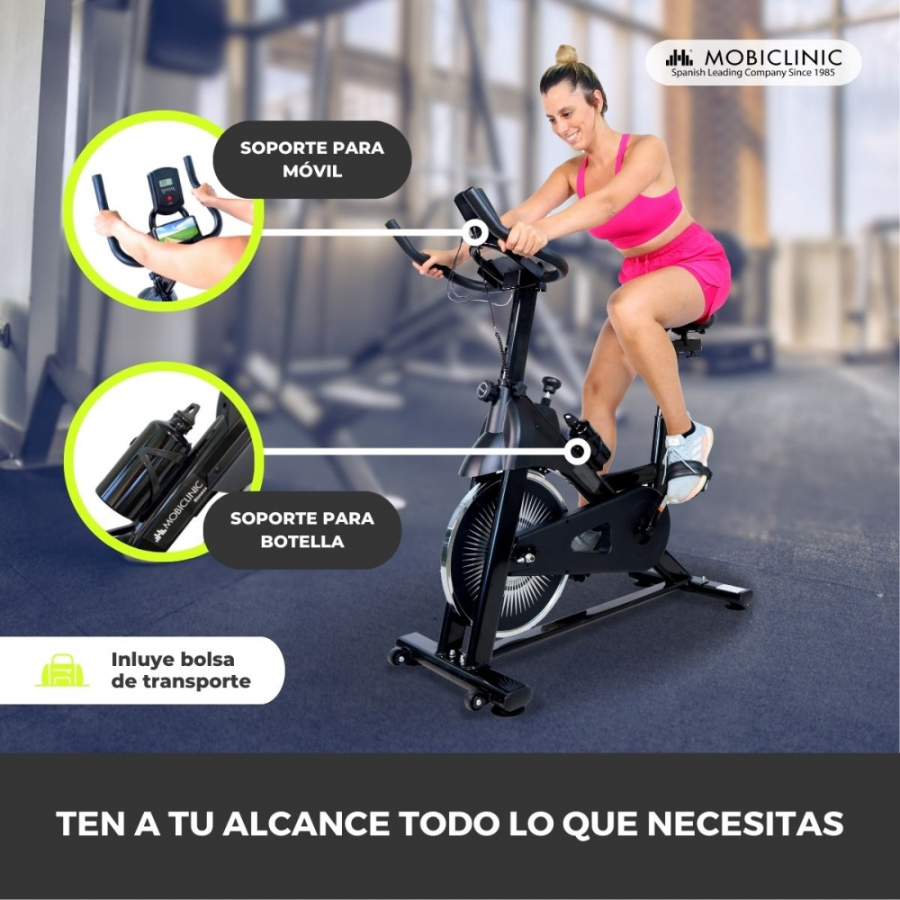 Una mujer en rosa hace ejercicio en una bicicleta estática negra. La imagen destaca características, como un soporte para teléfono y uno para botella.