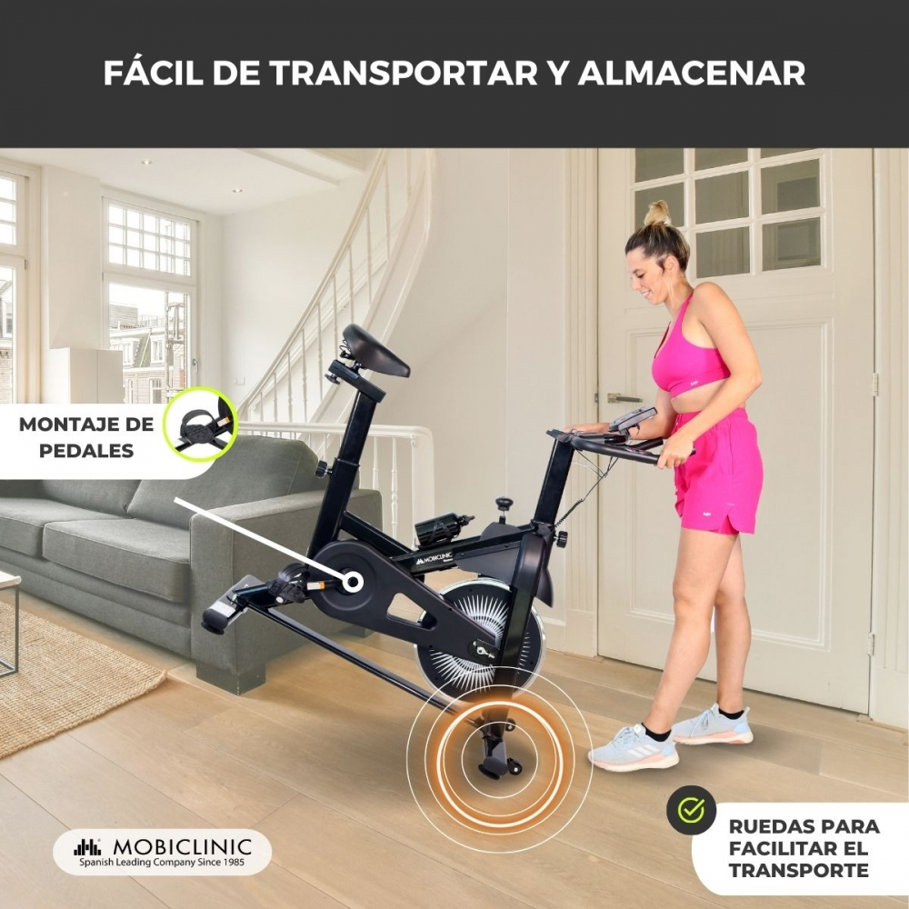 Mujer con ropa deportiva rosa y una bicicleta estática. La bicicleta tiene ruedas para facilitar el transporte. Sofá y puerta al fondo.