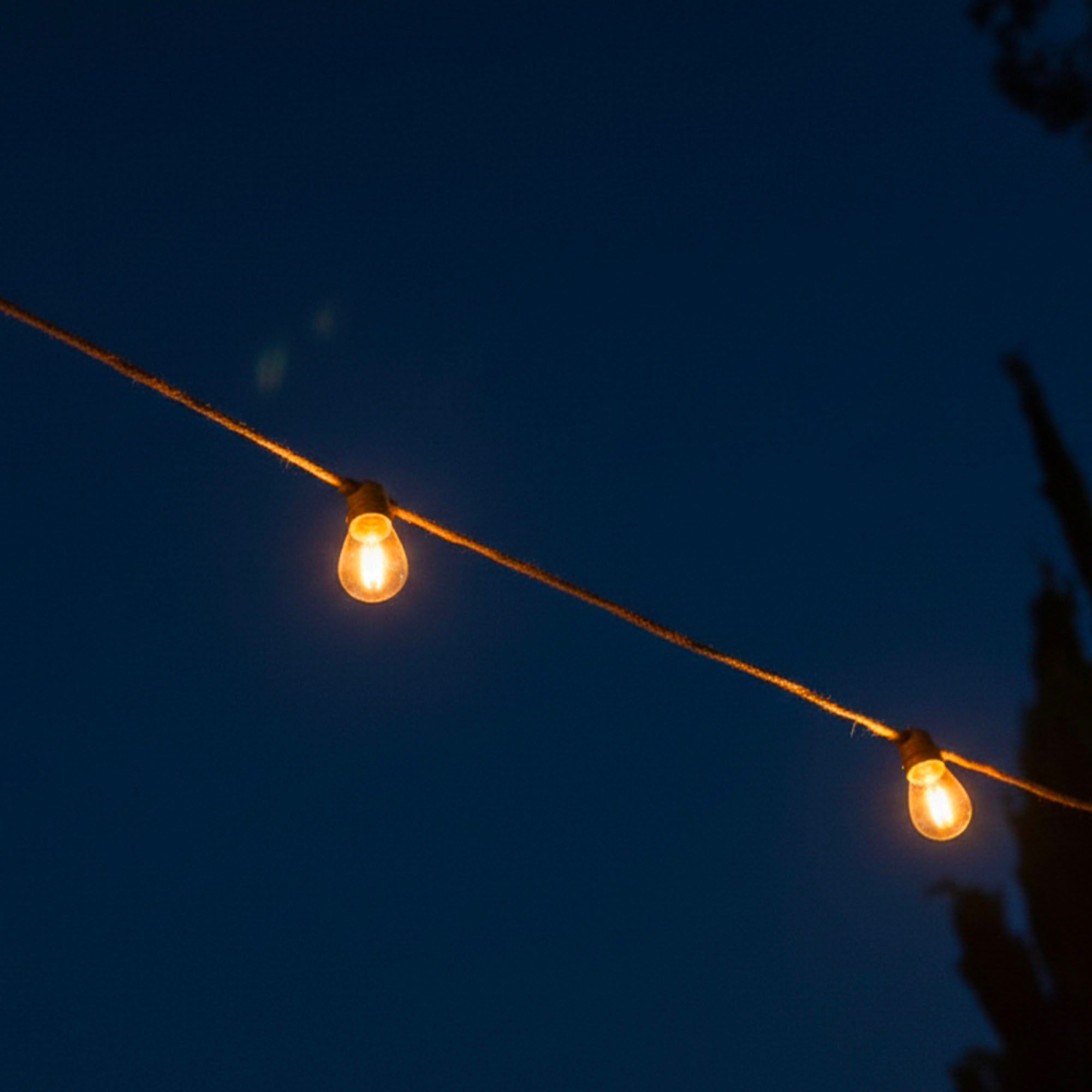 Lichterketten, zwei brennende Glühbirnen, vor dunklem blauem Himmel. Das Seil ist abgewinkelt, die Lichter leuchten.