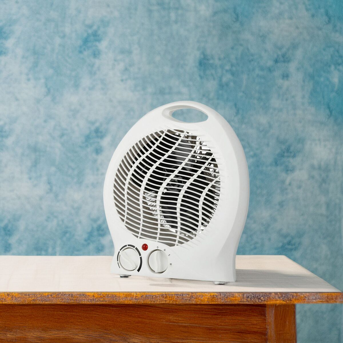 Un calefactor eléctrico blanco sobre una mesa de madera, contra una pared azul. Tiene dos diales y una luz roja.