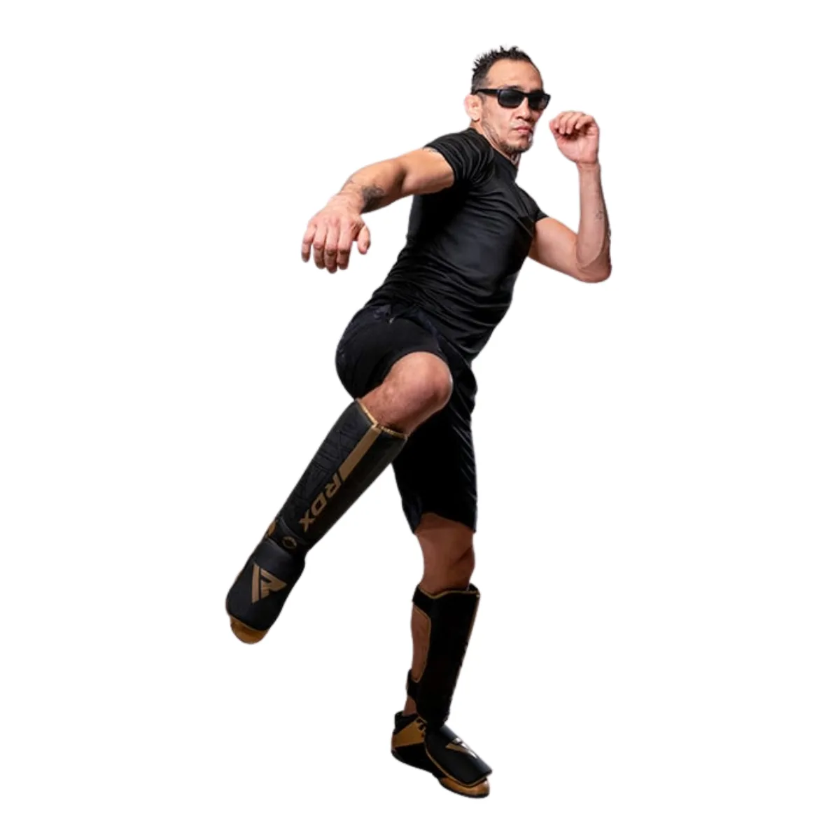 Ein Mann in schwarzer Sportkleidung und Sonnenbrille tritt. Er trägt Schienbeinschützer mit goldenen Akzenten. Weißer Hintergrund.