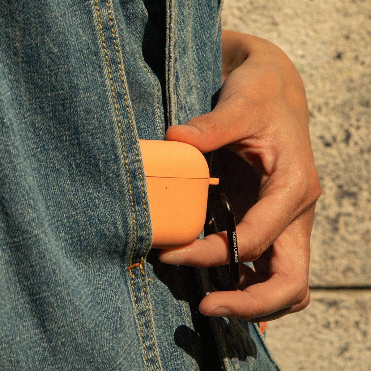 Una mano tiene una custodia arancione per AirPods agganciata a un moschettone nero, contro una giacca di jeans blu. Lo sfondo è un muro strutturato.