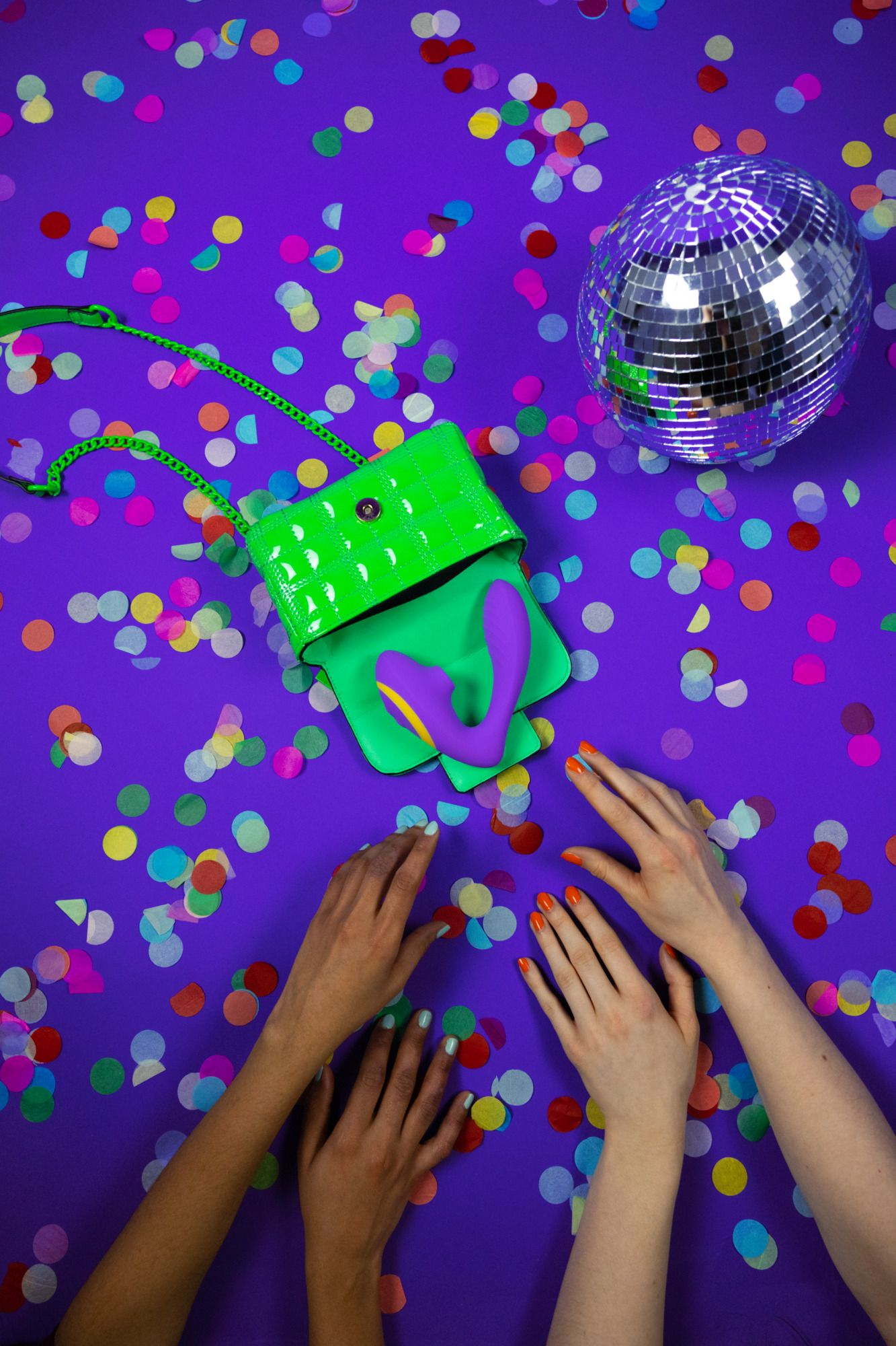 Un bolso verde con un juguete sexual morado sobre una superficie morada con confeti. Manos se acercan. Una bola de discoteca arriba.