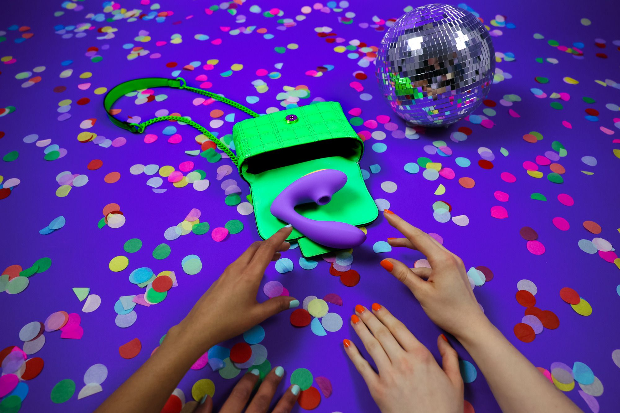 Vibrador morado, bolso verde, bola de discoteca, manos, confeti sobre fondo morado.