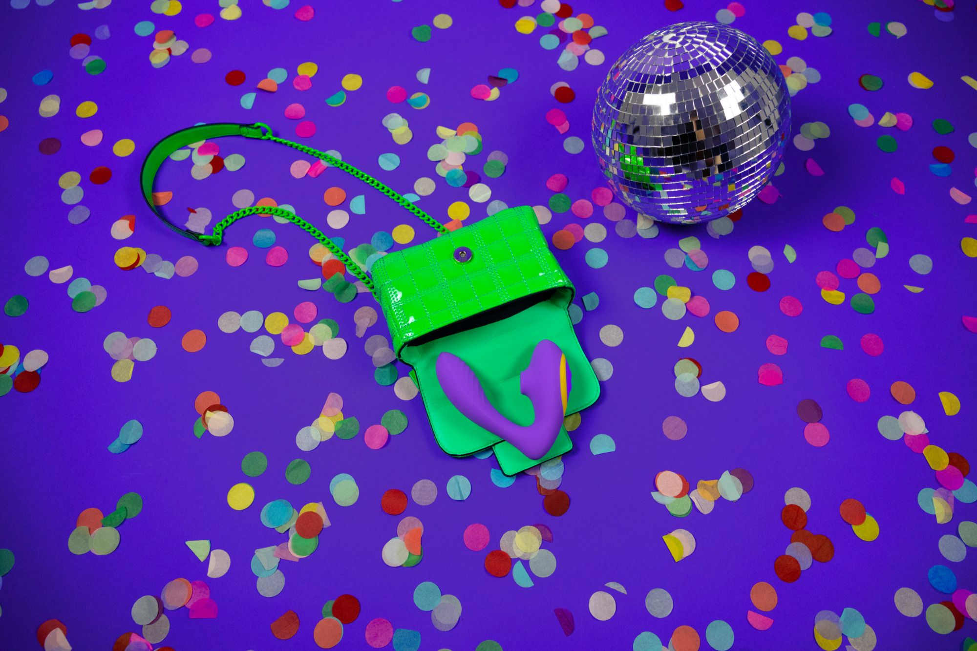 Un bolso verde con un juguete sexual morado está sobre una superficie morada salpicada de confeti, una bola de discoteca a la derecha.
