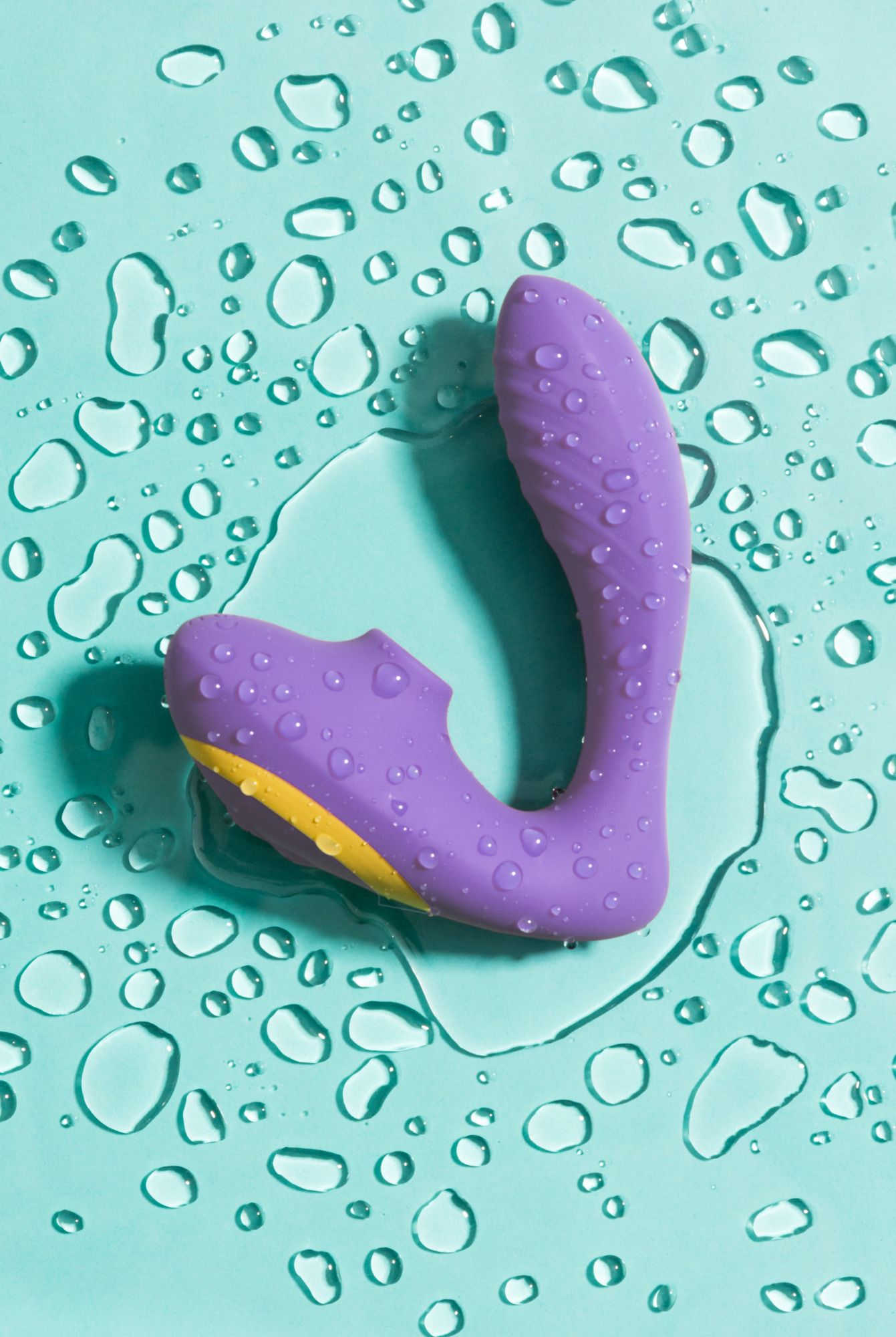 Un vibrador morado con una franja amarilla, cubierto de gotas de agua, descansa sobre una superficie turquesa con gotas de agua.