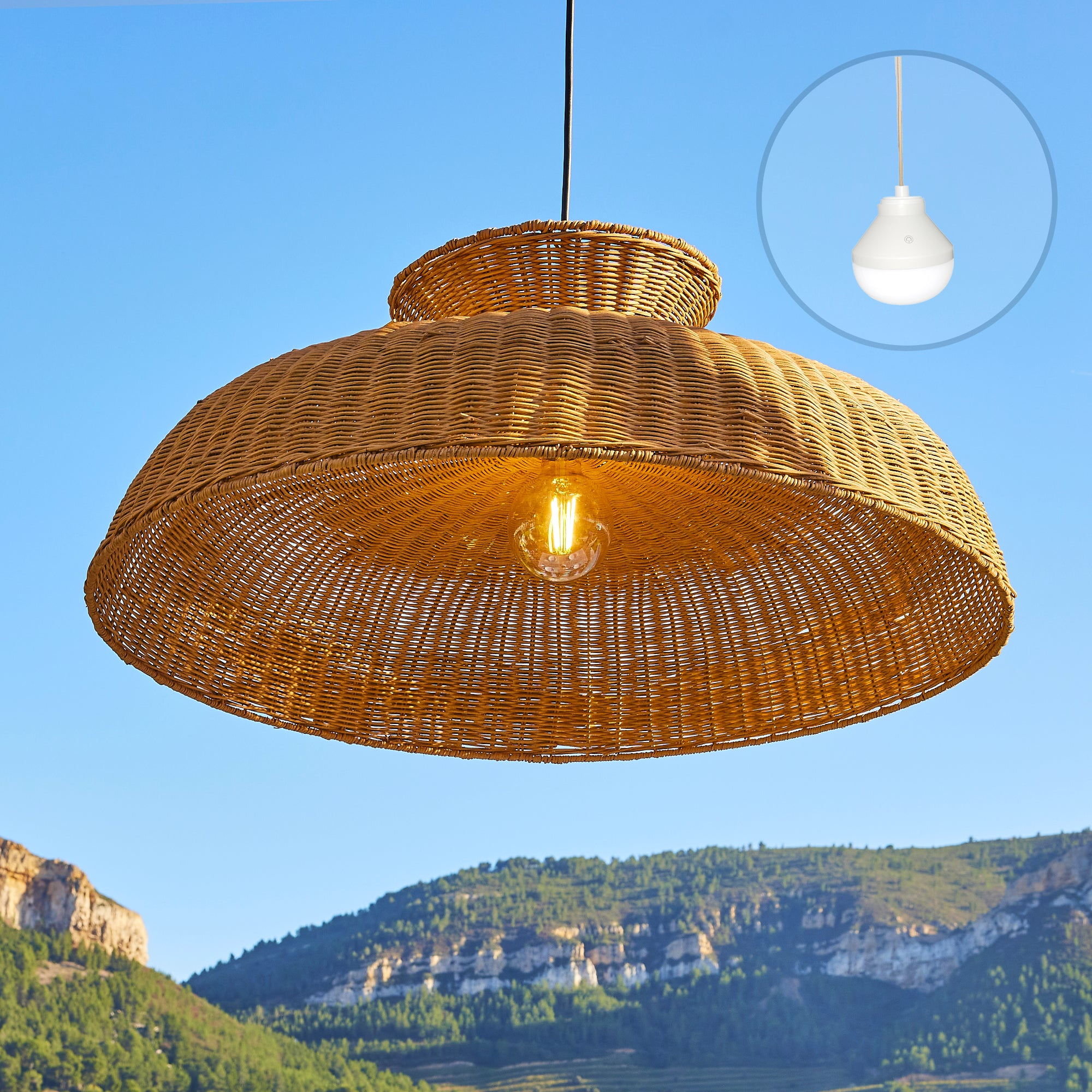 Ein gewebter Lampenschirm hängt, von innen beleuchtet, vor blauem Himmel. Berge sind im Hintergrund. Eine kleine Lampe ist eingesetzt.