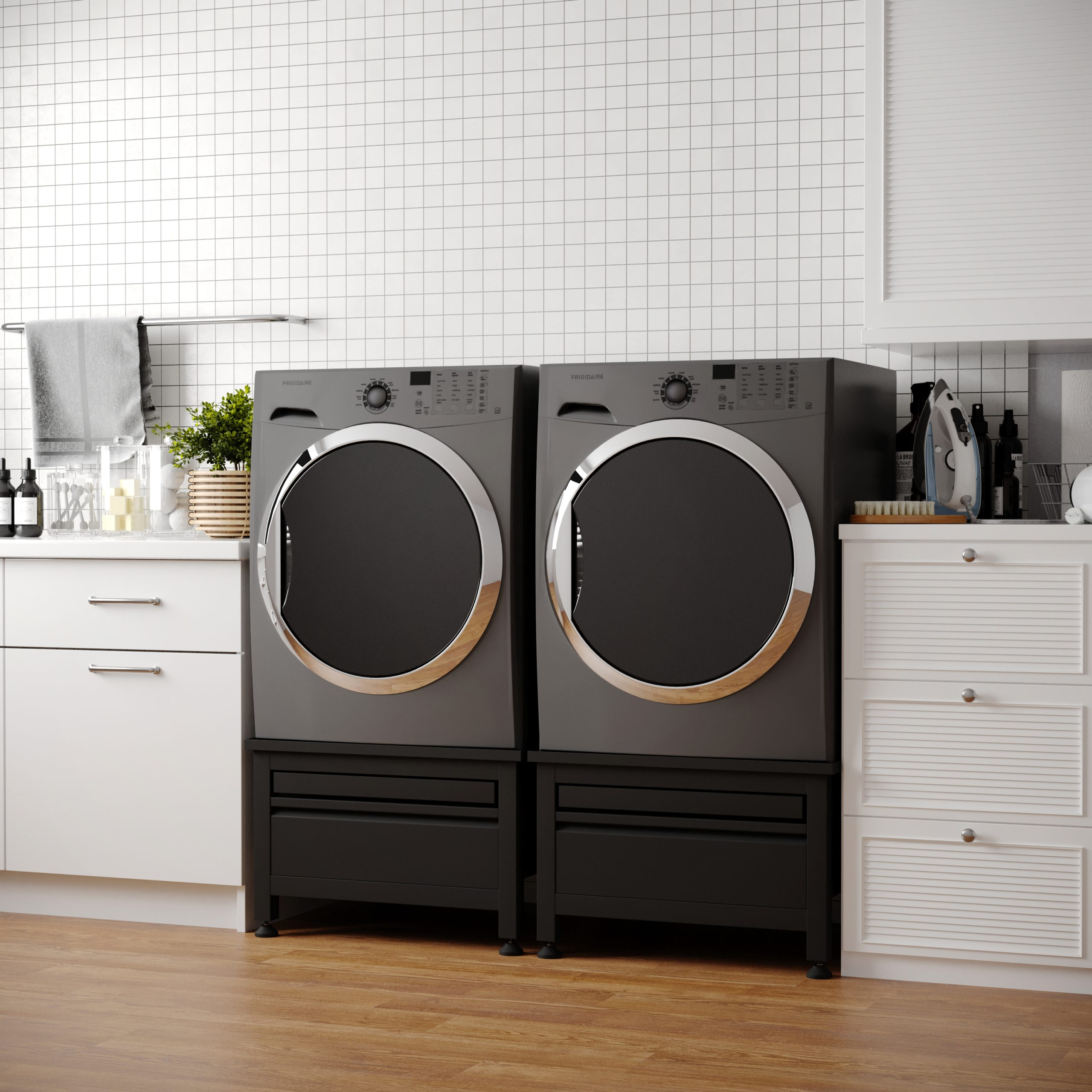 A laundry room with two gray Frigidaire dryers. White cabinets, iron, and a rack with towels are visible.