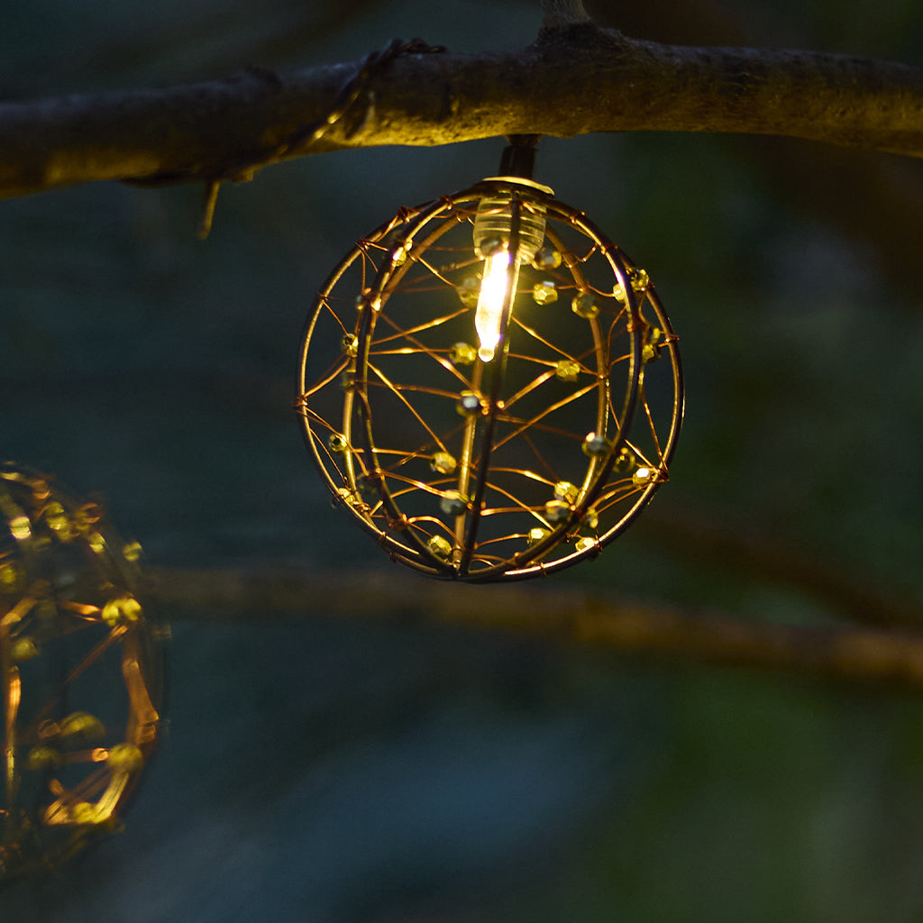 Eine leuchtende, kugelförmige Lampe mit Drahtrahmen-Design hängt an einem Ast. Kleine grüne Perlen verzieren die Struktur, mit warmem Licht.