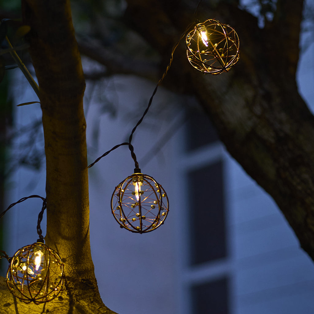 Lichterketten mit kugelförmigen Metallkäfigen hängen an Ästen. Warme gelbe Lichter leuchten am Abend. Im Hintergrund ist ein Gebäude.