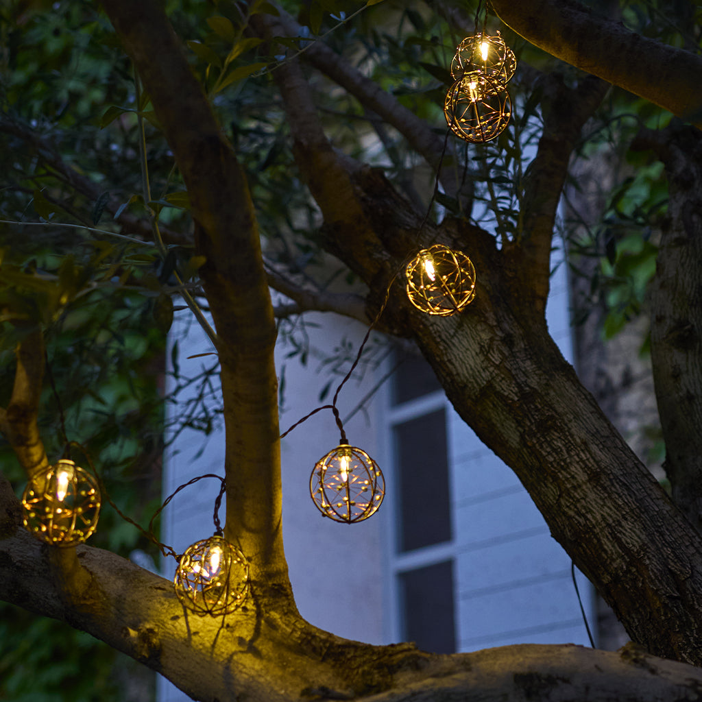 Lichterketten mit kugelförmigen Metallkäfigen hängen an Ästen. Die Lichter sind warm, und im Hintergrund ist ein Haus zu sehen.
