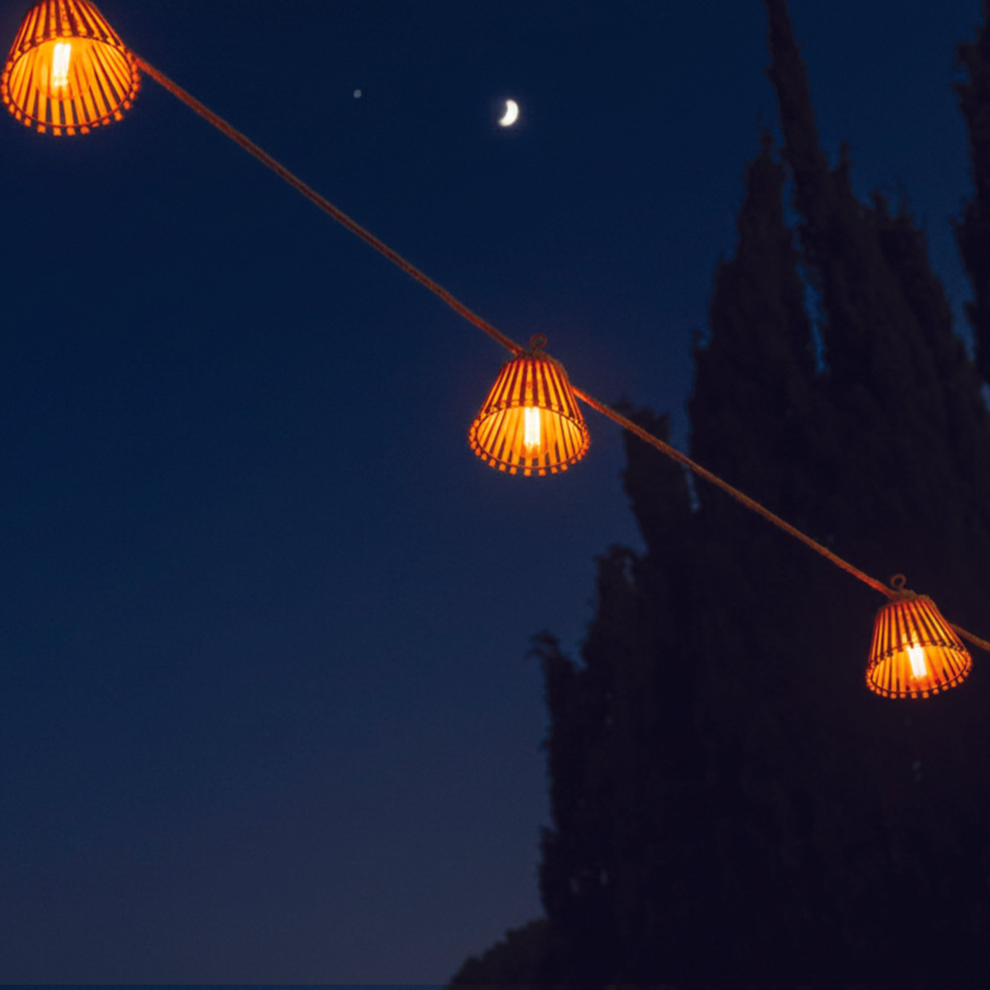 Nachtszenen mit Lichterketten, einem Halbmond und Bäumen als Silhouette vor dunkelblauem Himmel.