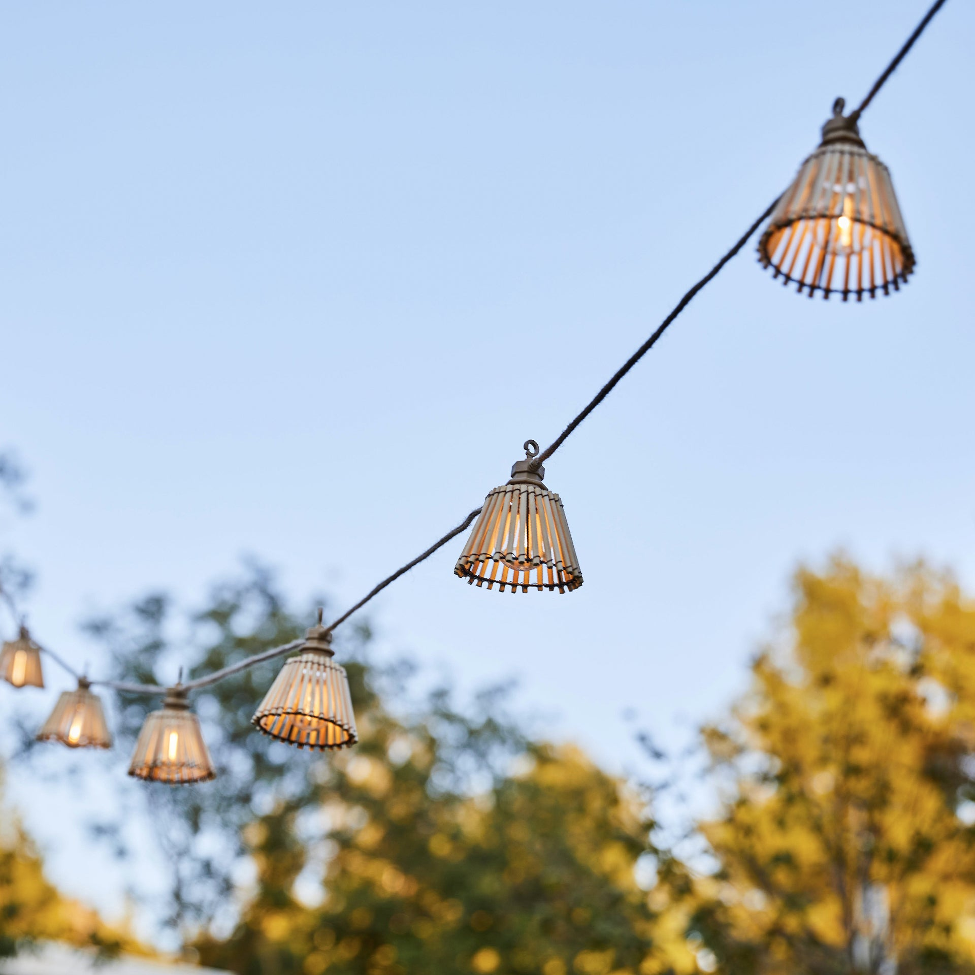 Lichterketten mit Bambus-Lampenschirmen hängen vor blauem Himmel, mit verschwommenen Herbstbäumen im Hintergrund.