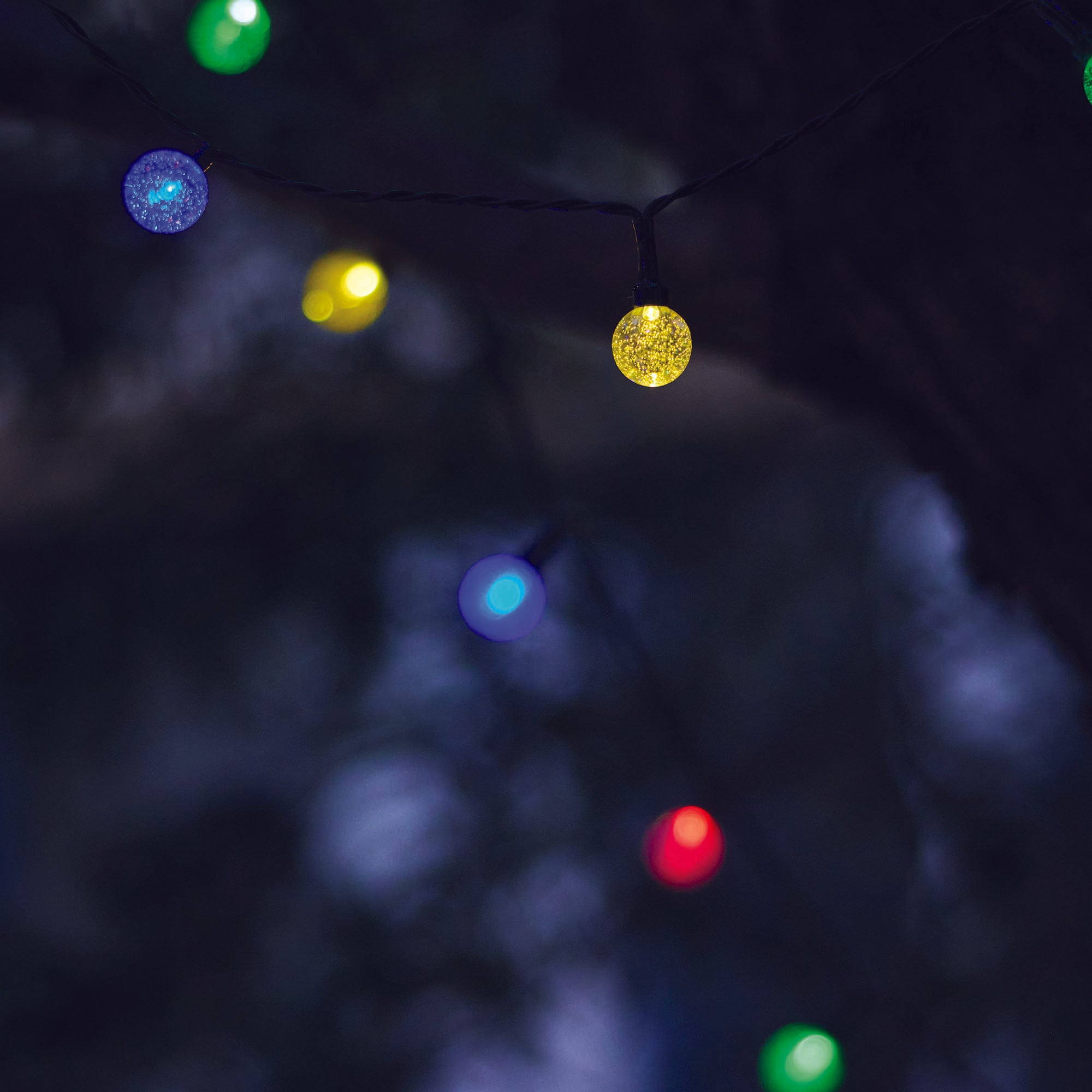 Eine Lichterkette mit bunten, runden Lichtern, die vor einem dunklen, verschwommenen Hintergrund hängen. Die Lichter sind grün, gelb, blau und rot.