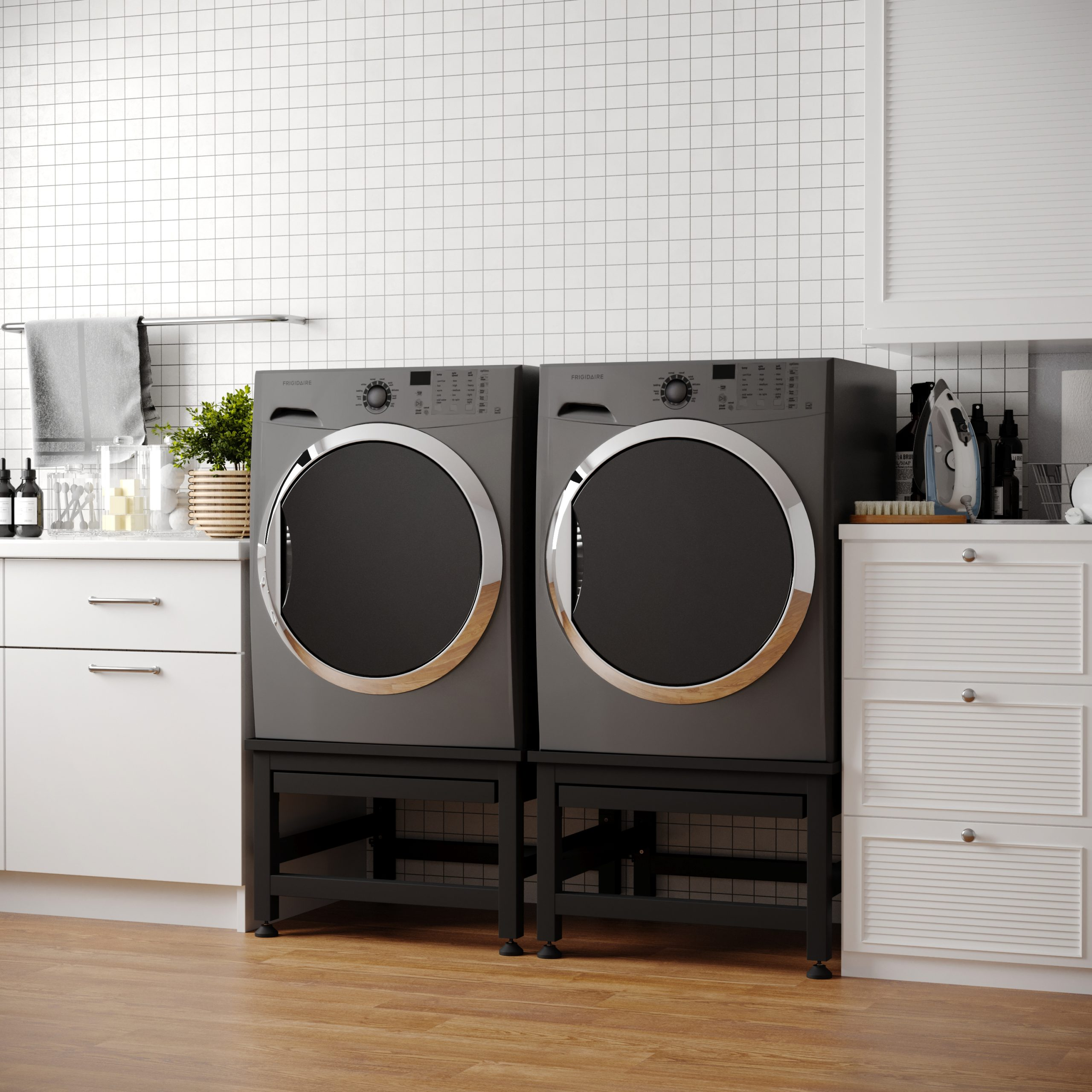 Twee grijze wasmachines staan naast elkaar op zwarte standaards, in een wasruimte met witte kasten.