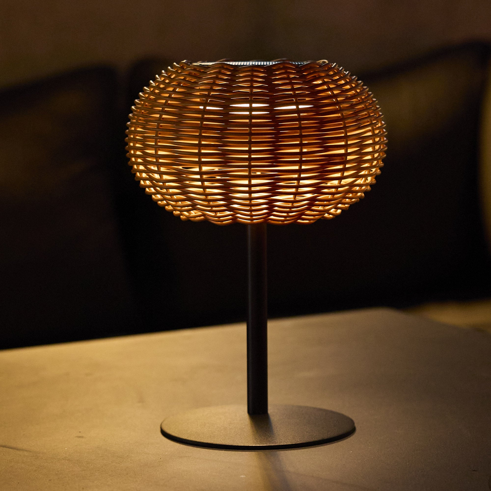 Eine gewebte Kugellampe auf einem Ständer. Die Lampe leuchtet und wirft einen warmen Schein. Dunkler Hintergrund.