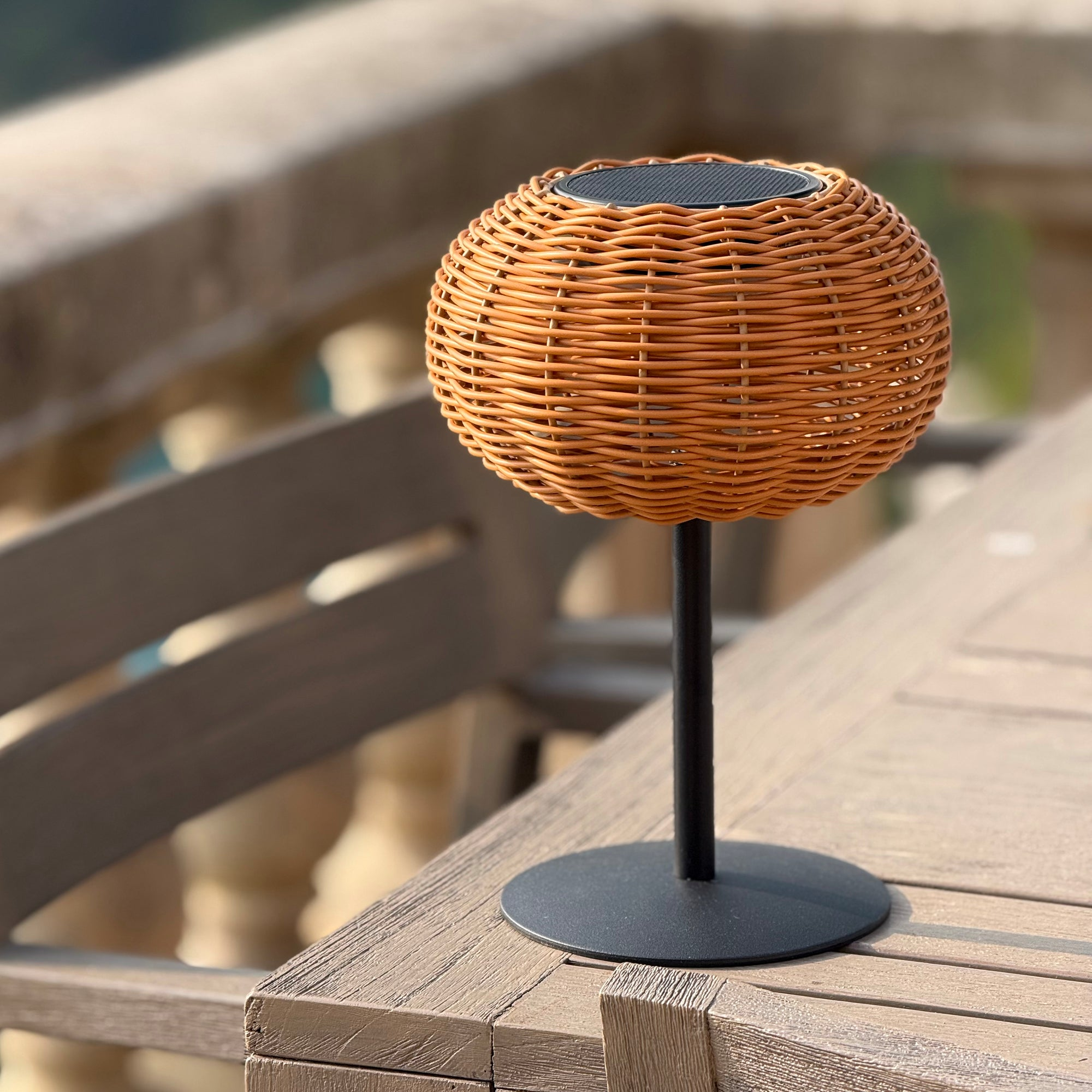 Eine gewebte, kugelförmige Lampe steht auf einem Holztisch. Die Lampe hat einen schwarzen Fuß und Stiel. Der Hintergrund ist verschwommen.