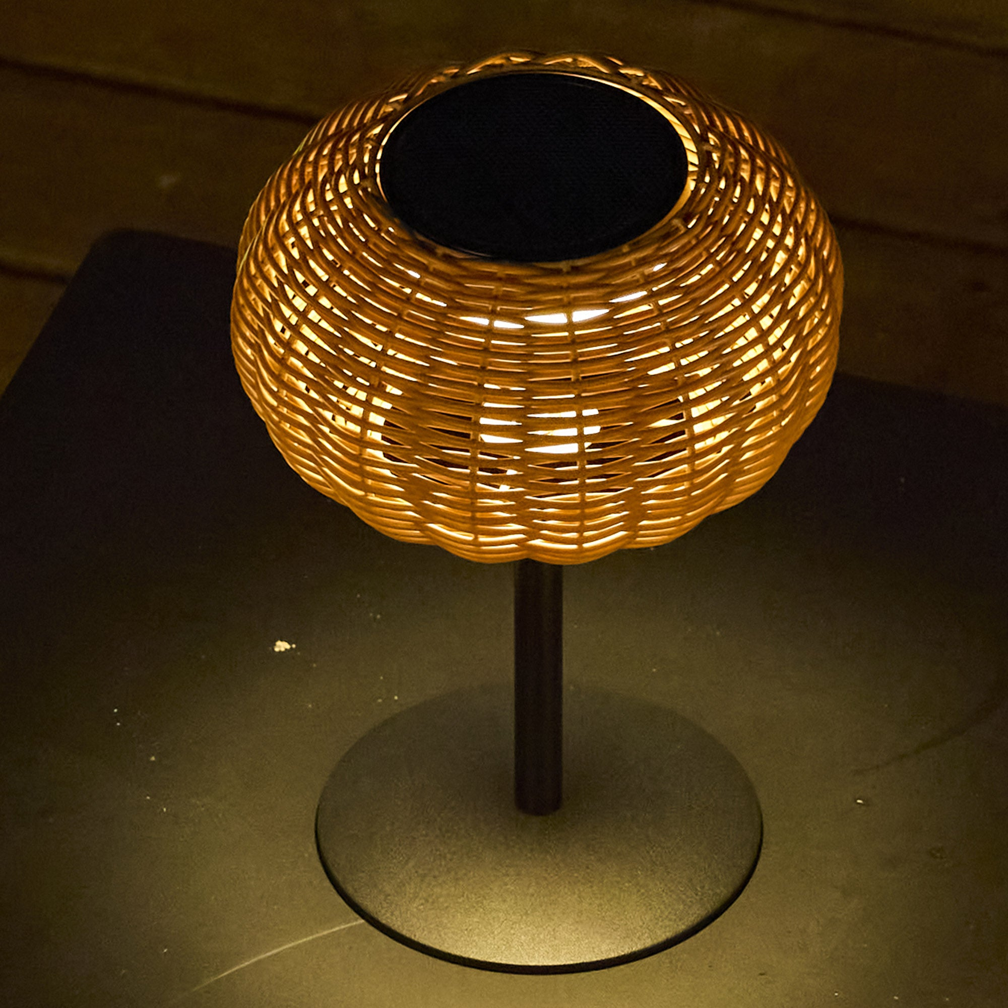 Eine gewebte Rattanlampe mit schwarzer Oberseite. Sie steht auf einem dunklen Ständer auf einem dunklen Tisch.