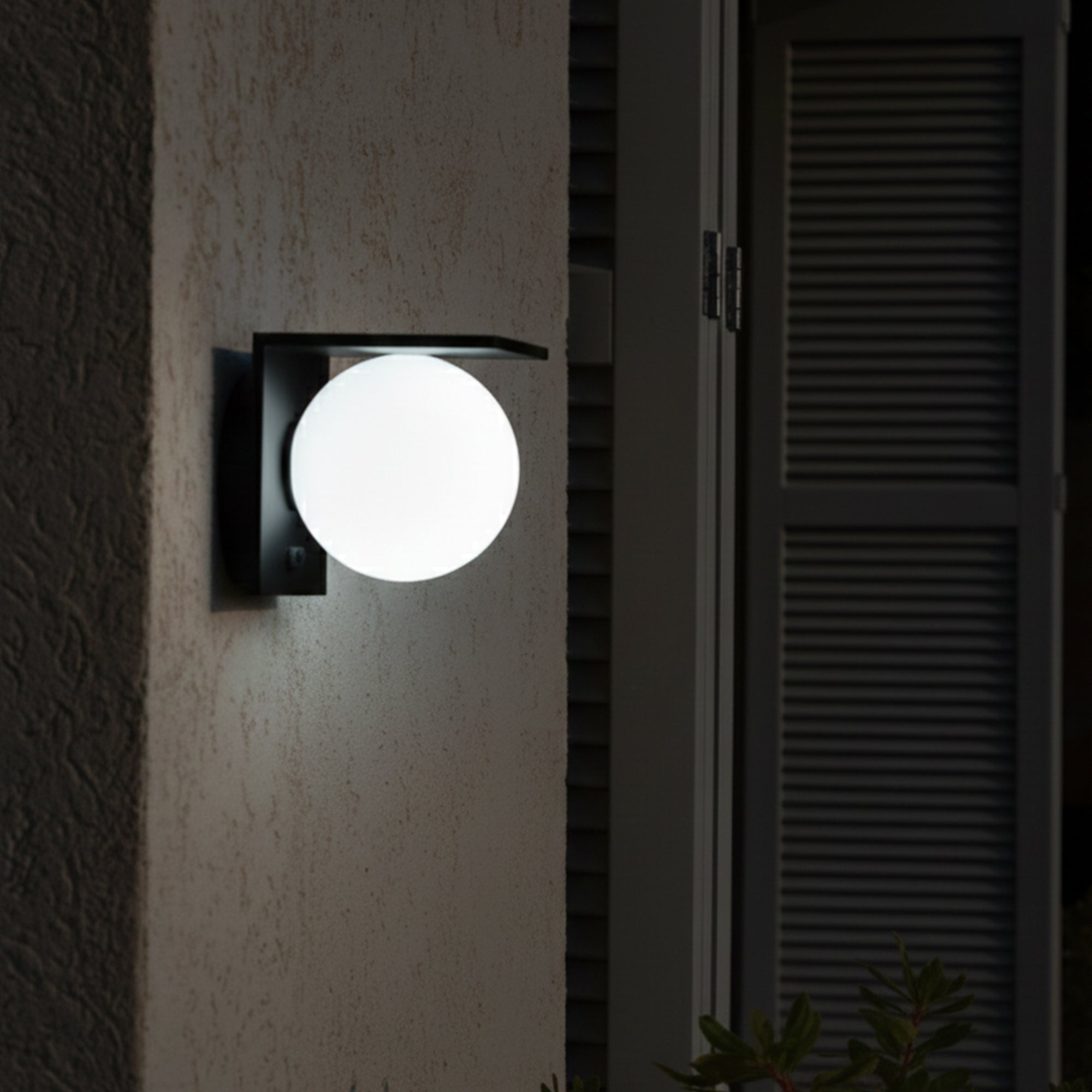 Eine runde, weiße Außenlampe mit schwarzem Halter ist an einer strukturierten Wand neben einer Fensterläden-Tür montiert.