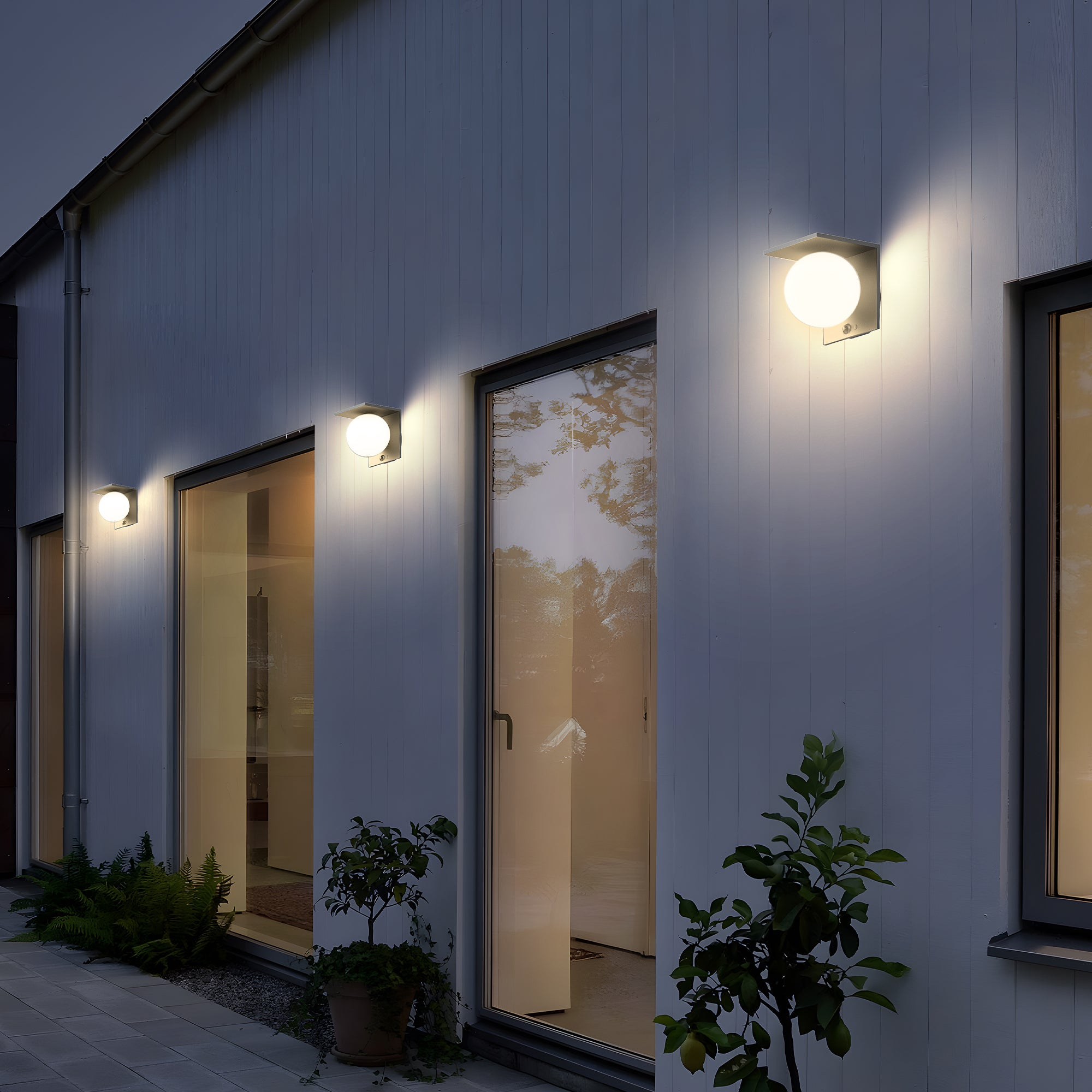Außenaufnahme eines modernen Gebäudes in der Abenddämmerung. Wandleuchten beleuchten die Fassade, Pflanzen in Töpfen.