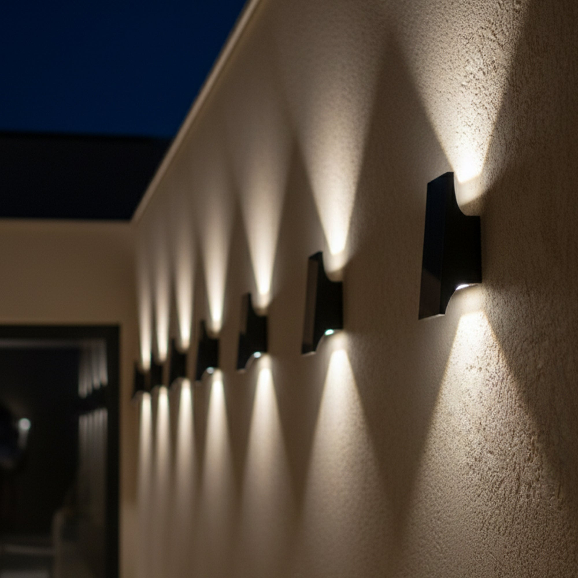 Außenwand mit mehreren schwarzen Wandleuchten. Jedes Licht scheint nach unten und beleuchtet die strukturierte Wand. Dunkler Himmel im Hintergrund.