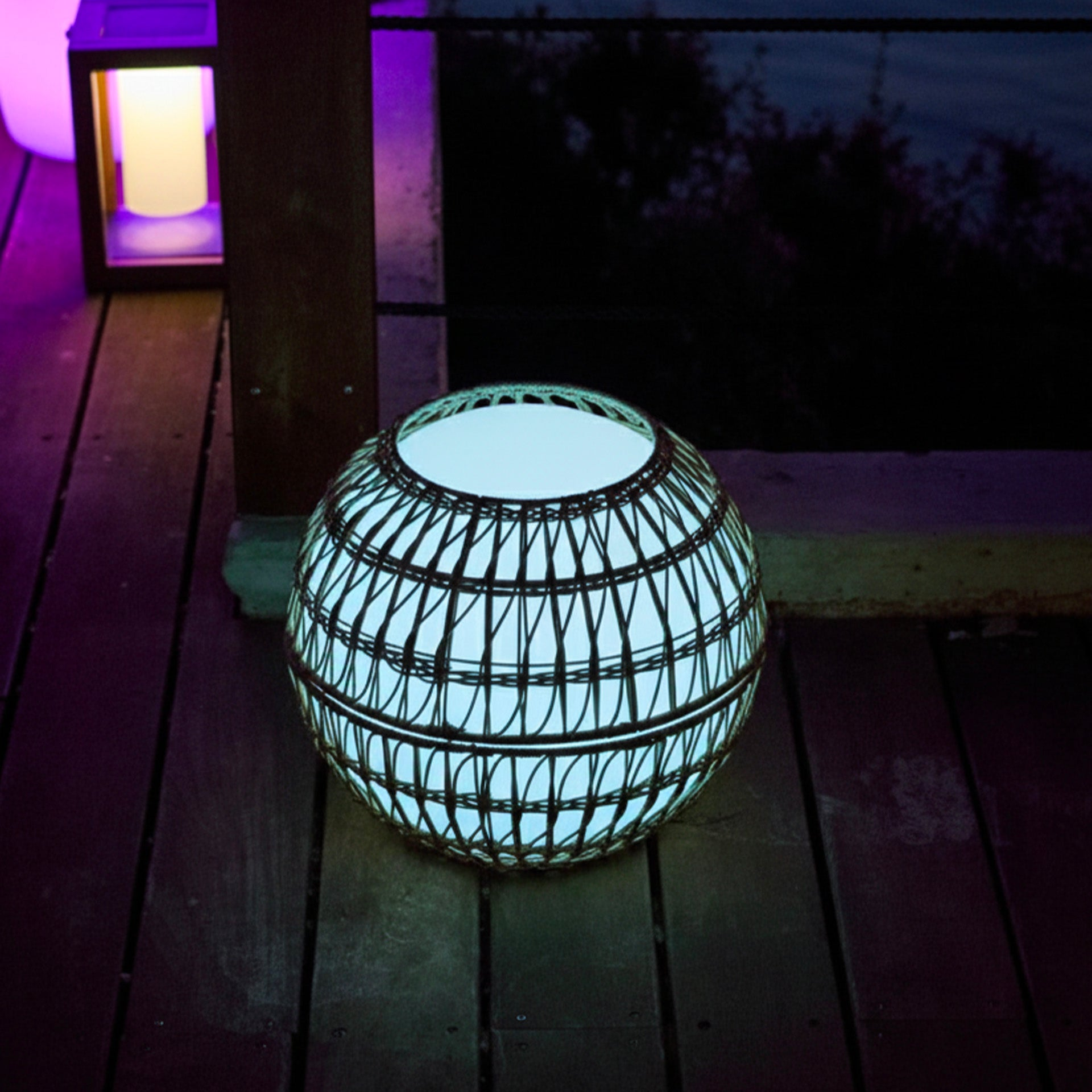 Eine leuchtende kugelförmige Lampe aus gewebtem Material steht auf einem Holzdeck. Eine Laterne und dunkler Hintergrund sind sichtbar.