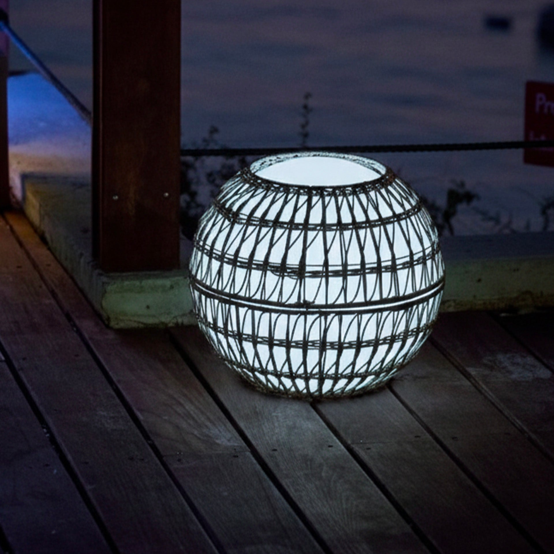 Eine leuchtende kugelförmige Lampe aus gewebtem Material steht auf einem Holzdeck. Der Hintergrund zeigt einen dunklen Himmel und eine Holzkonstruktion.