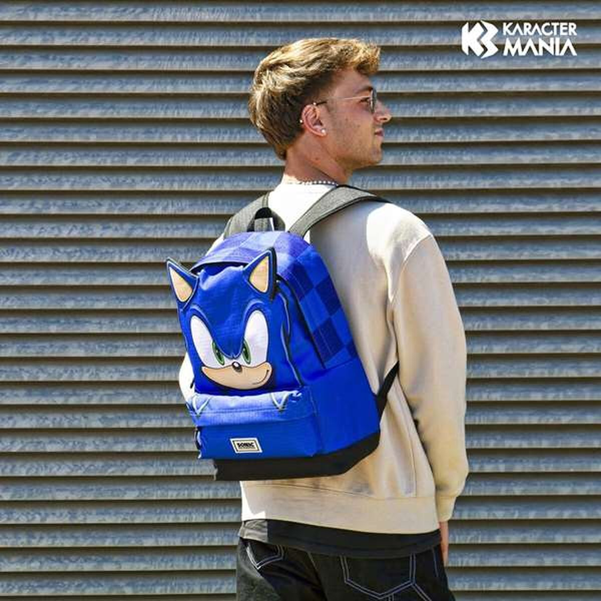 Un uomo con uno zaino con la faccia di Sonic the Hedgehog. Indossa occhiali da sole e un maglione color crema, in piedi davanti a un muro.