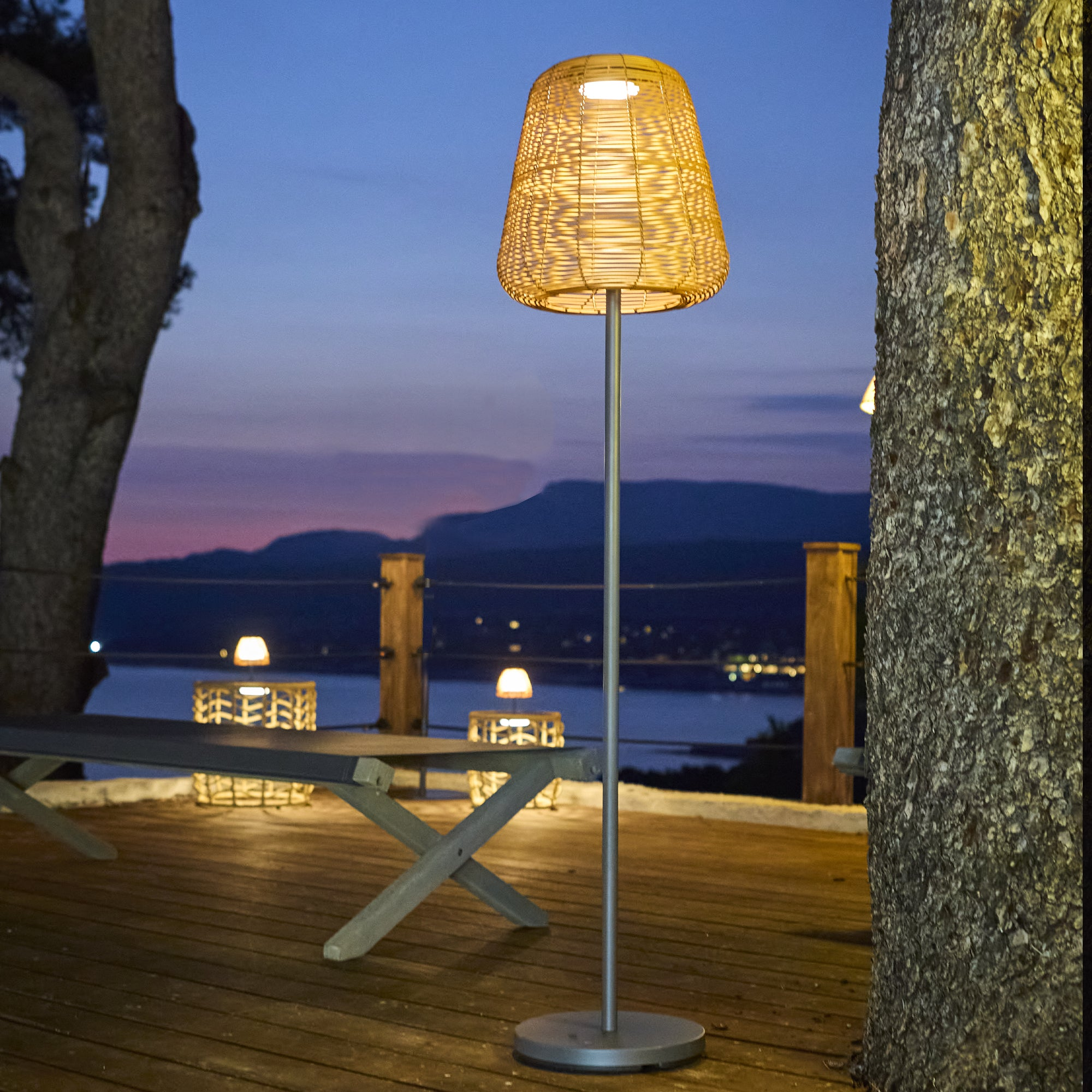 Eine beleuchtete gewebte Lampe steht auf einer Holzterrasse in der Dämmerung, mit Tischen und Blick auf das Meer und die Berge.