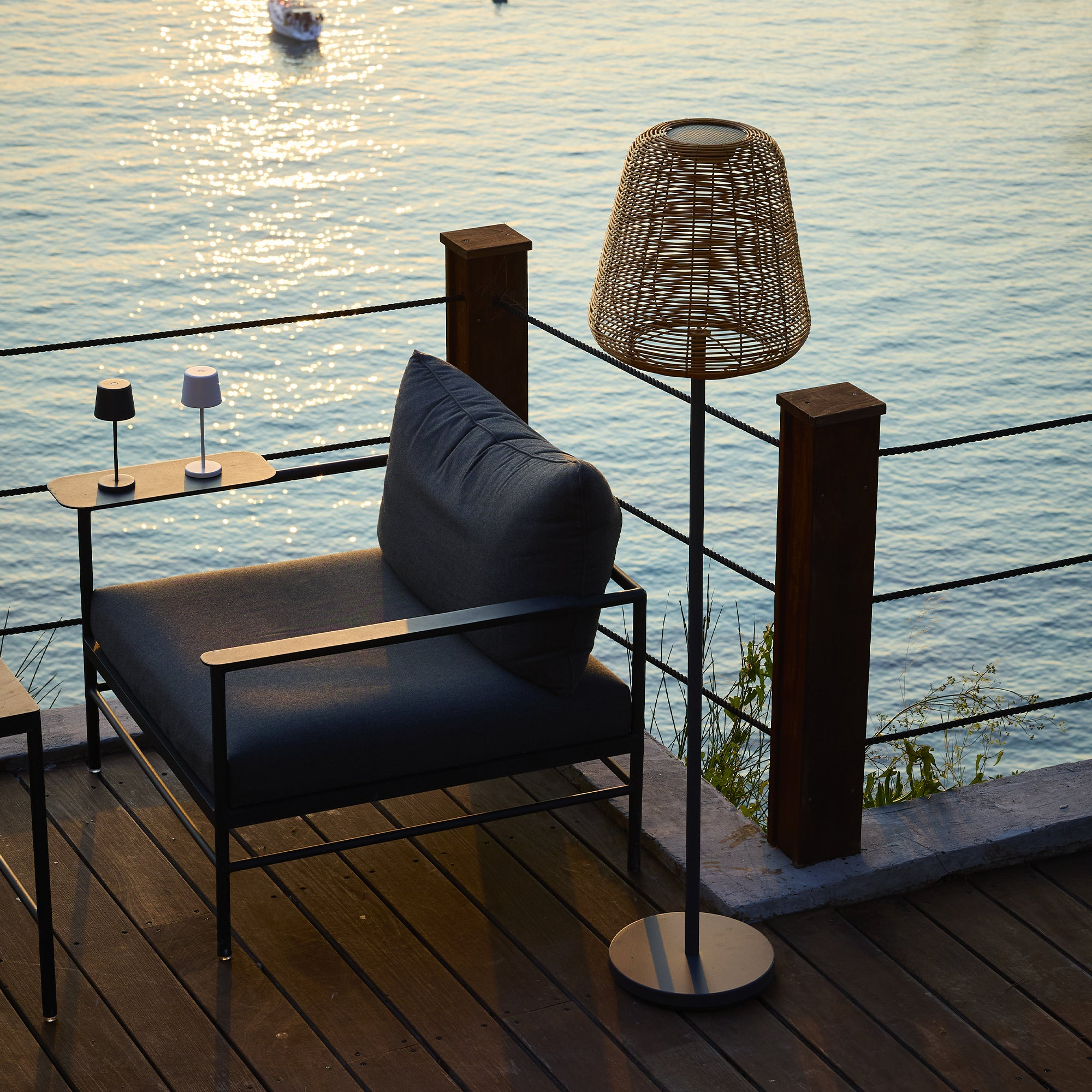 Eine Terrasse mit Stuhl, Lampen und Wasser im Hintergrund. Ein Boot ist zu sehen. Die Szene wird von der untergehenden Sonne beleuchtet.