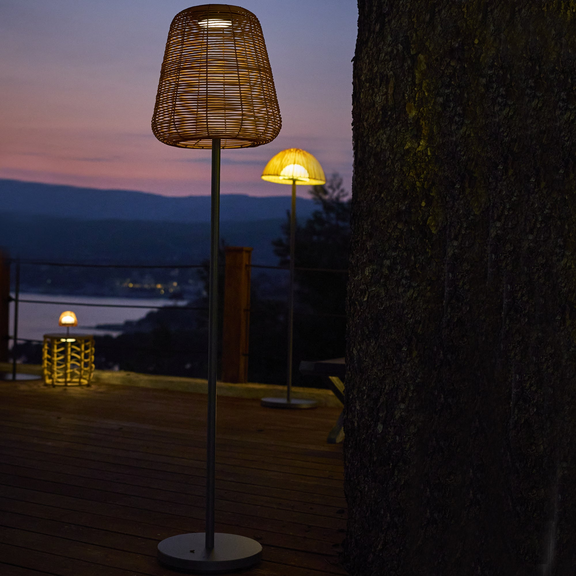 Abendliche Szene mit beleuchteten Lampen auf einem Deck. Korblampen stehen vor einem dunklen Himmel und Bergen.