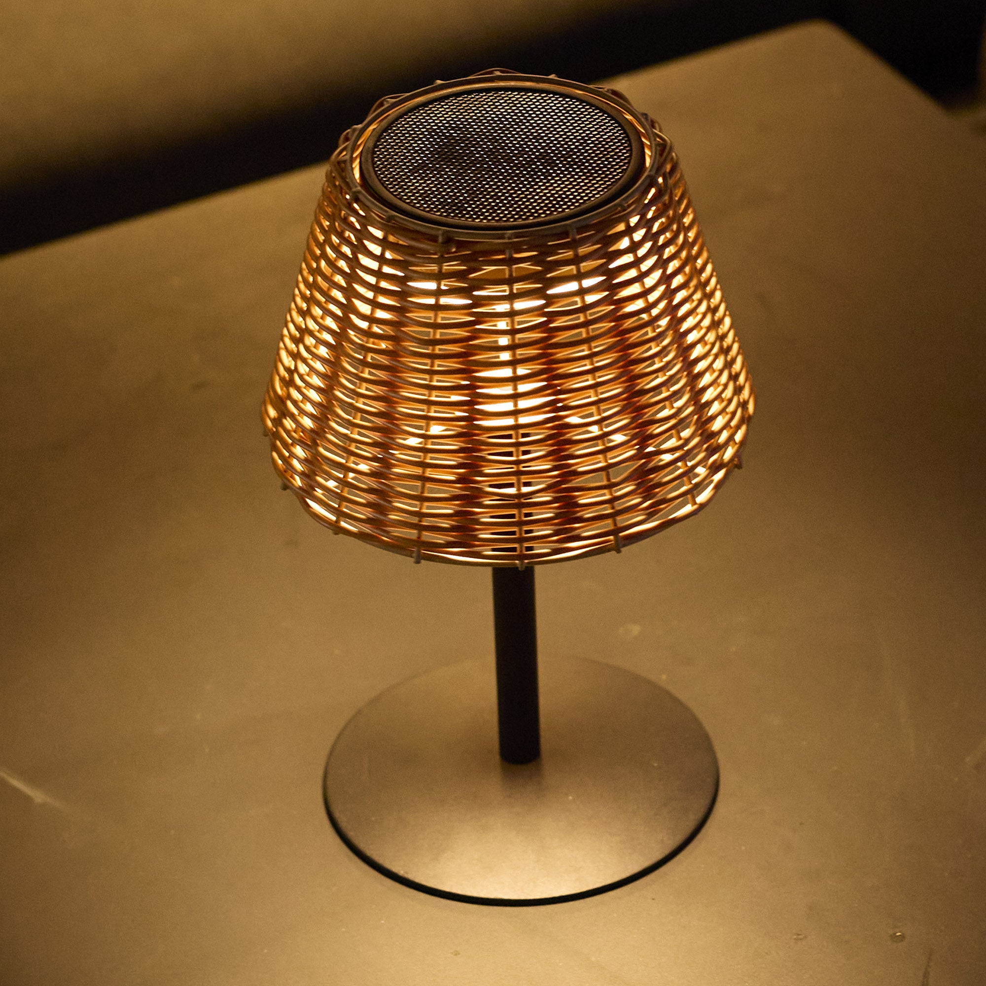 Eine gewebte Lampe mit zylindrischem Schirm, von innen beleuchtet, steht auf einem dunklen, kreisförmigen Sockel.