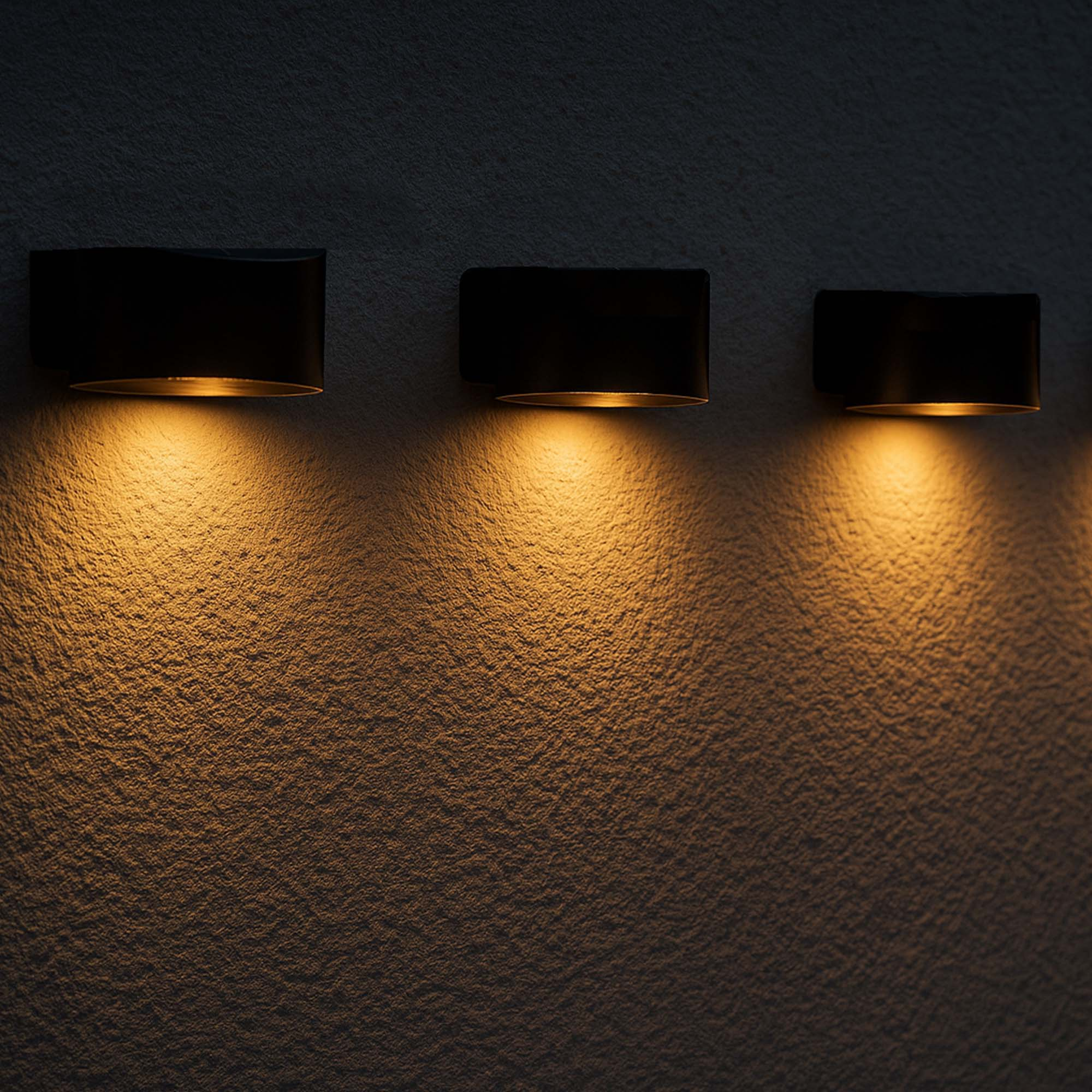 Drei schwarze Wandlampen beleuchten eine strukturierte Wand mit warmem Licht und erzeugen einen Kontrast.