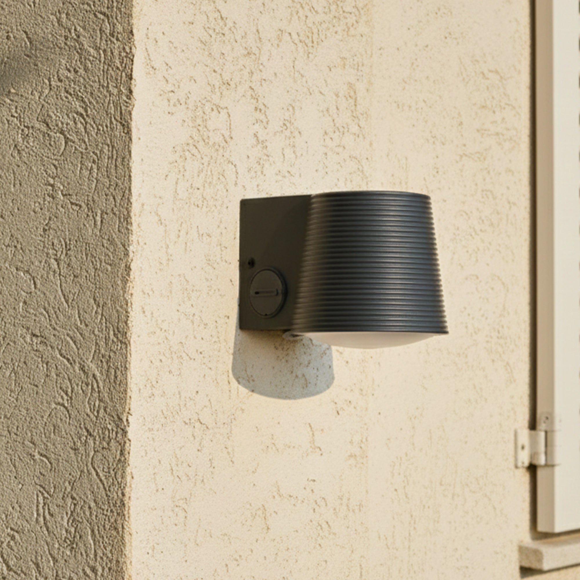 Eine dunkelgraue Außenlampe ist an einer strukturierten beigefarbenen Wand befestigt. Rechts ist ein weißer Fensterladen zu sehen.