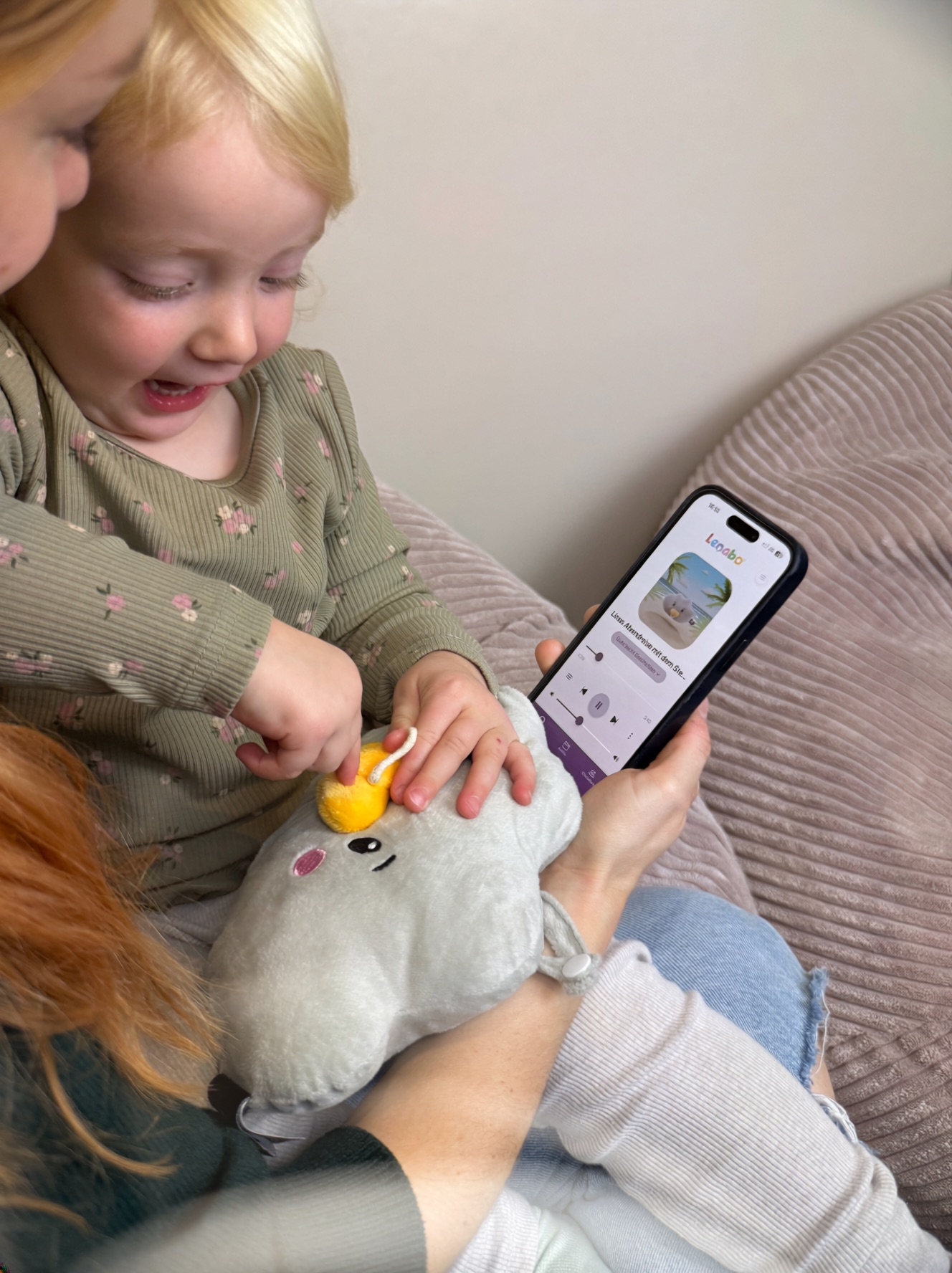 Ein Kind spielt mit einem Spielzeug, während eine Person ein Telefon mit einer App hält. Das Kind ist blond und trägt Grün.