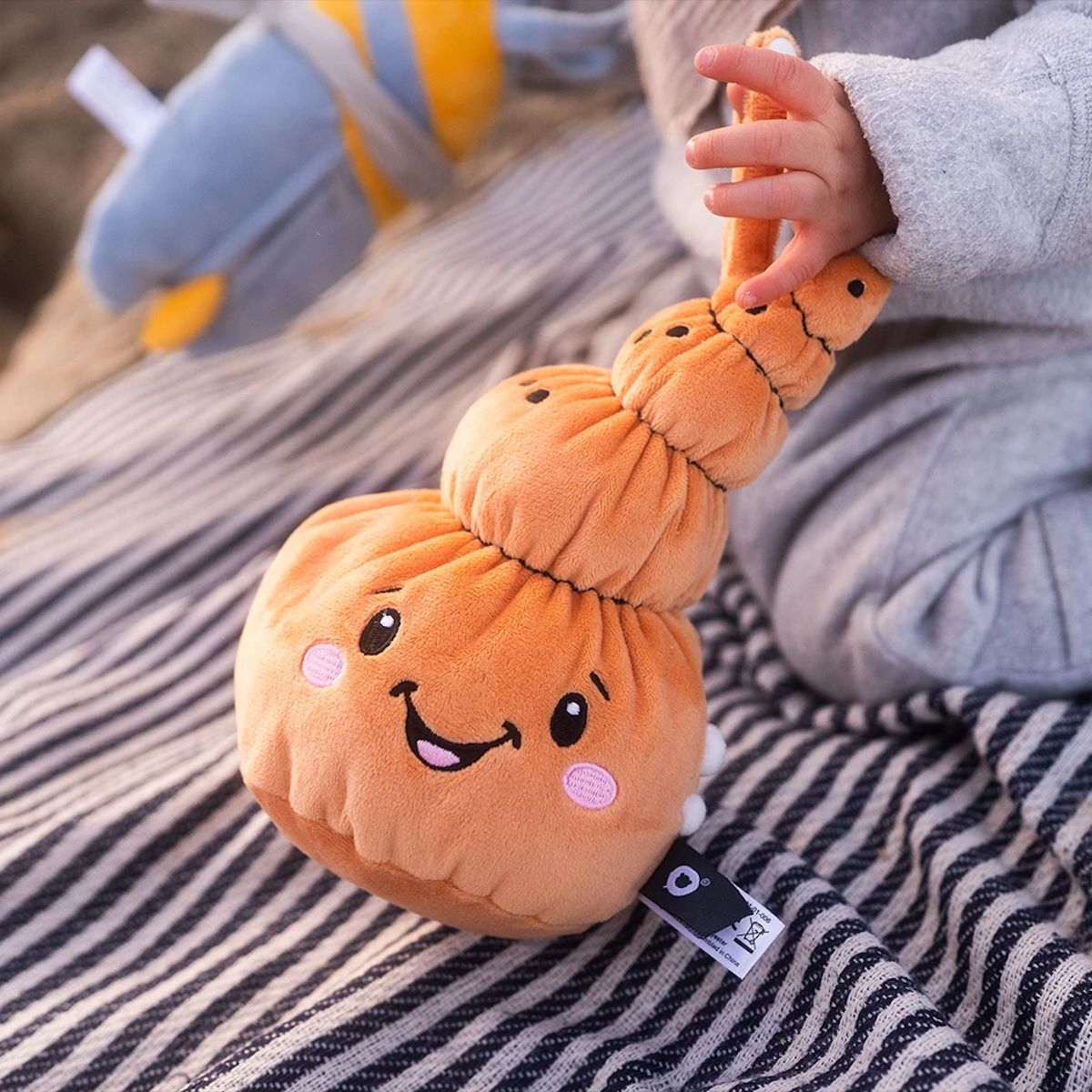 Die Hand eines Kindes hält ein oranges Plüschtier in Schneckenform. Das Spielzeug hat ein lächelndes Gesicht. Es liegt auf einer gestreiften Decke.