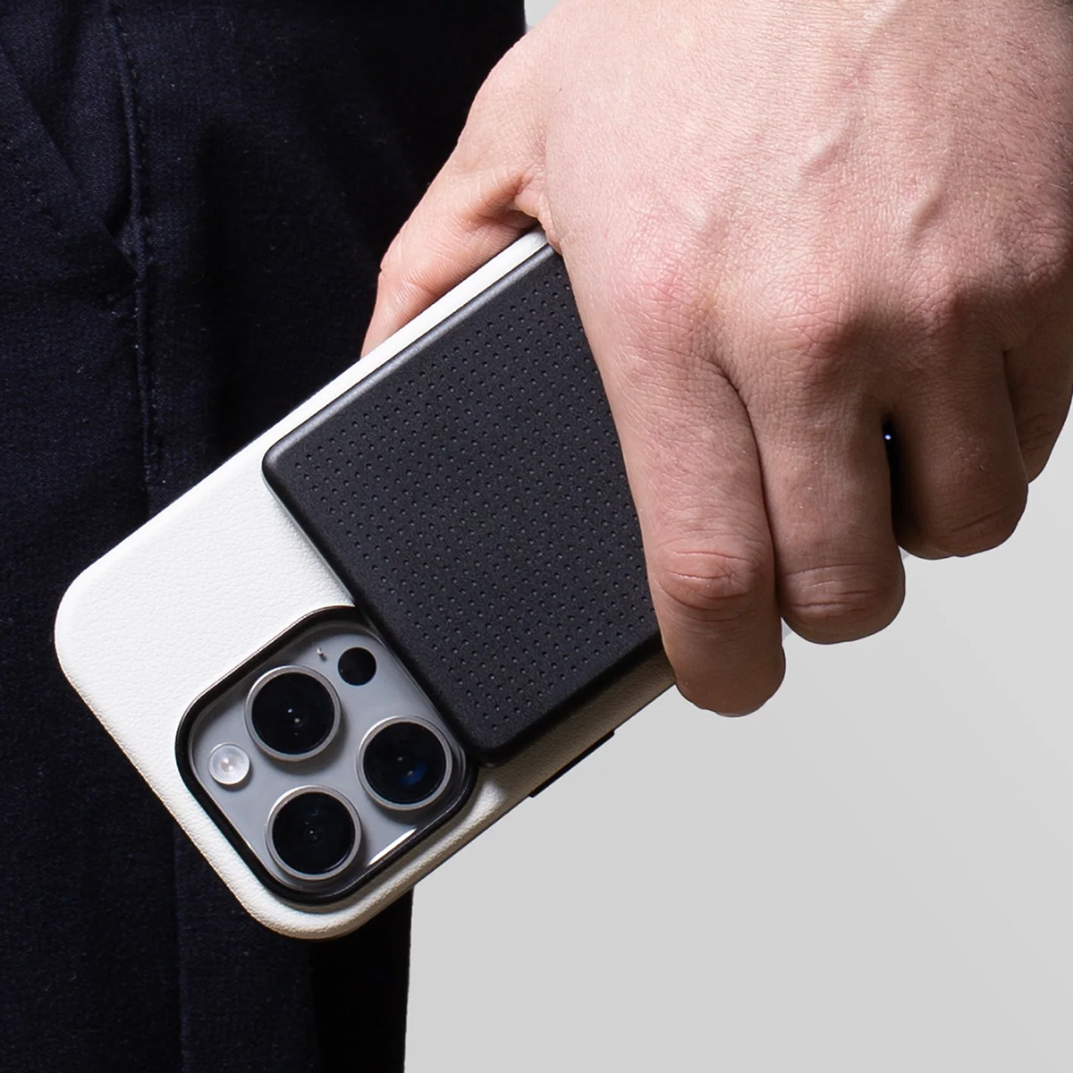 Une main tenant un étui de téléphone blanc et noir avec un téléphone. Le téléphone a trois caméras. La personne porte un pantalon noir.