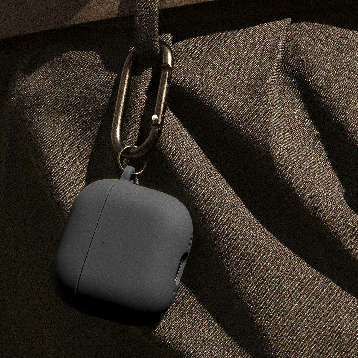 Ein dunkelgraues AirPods-Etui ist an einem metallischen Karabiner befestigt. Der Karabiner hängt an einem braunen Stoff.