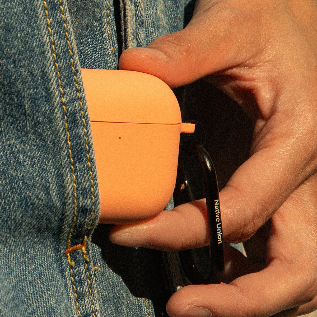 Una mano tiene una custodia arancione per AirPods agganciata a una tasca di jeans. La custodia ha un moschettone nero con 'Native Union' stampato.