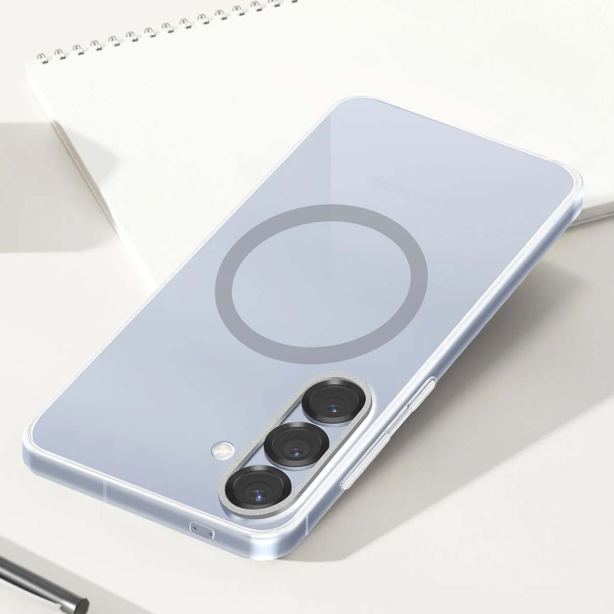 Una custodia trasparente con un design circolare, che mostra una fotocamera a tripla lente. Poggia su una superficie bianca accanto a un taccuino.