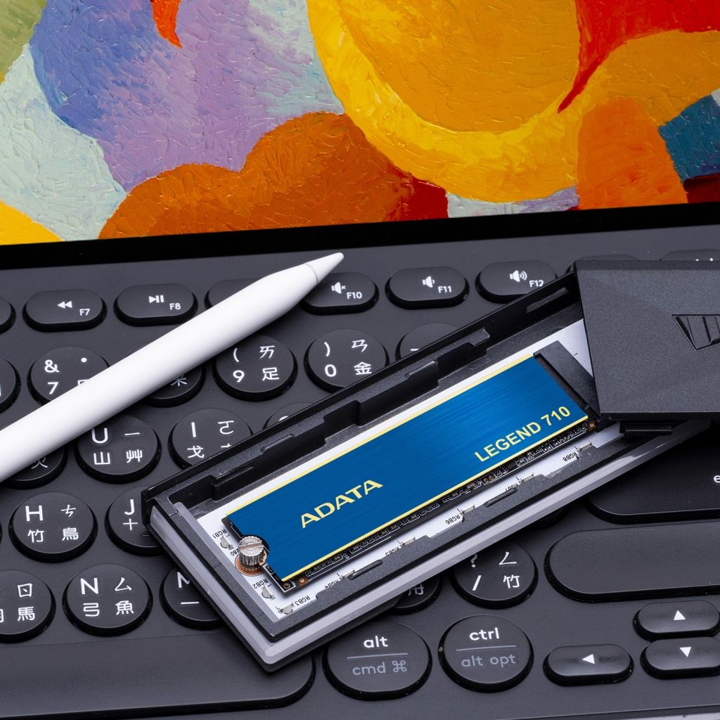 Klawiatura komputera z białym rysikiem, dyskiem SSD ADATA Legend 710 i kolorowym abstrakcyjnym obrazem w tle.