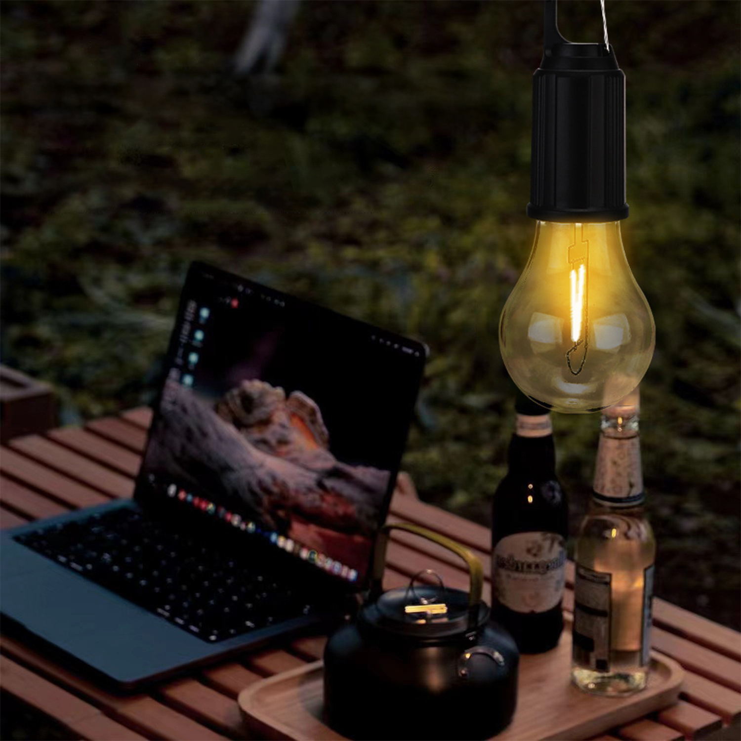 Una lampadina accesa è appesa sopra un laptop, bottiglie e un bollitore su un tavolo di legno. Lo sfondo è un ambiente esterno verde e sfocato.