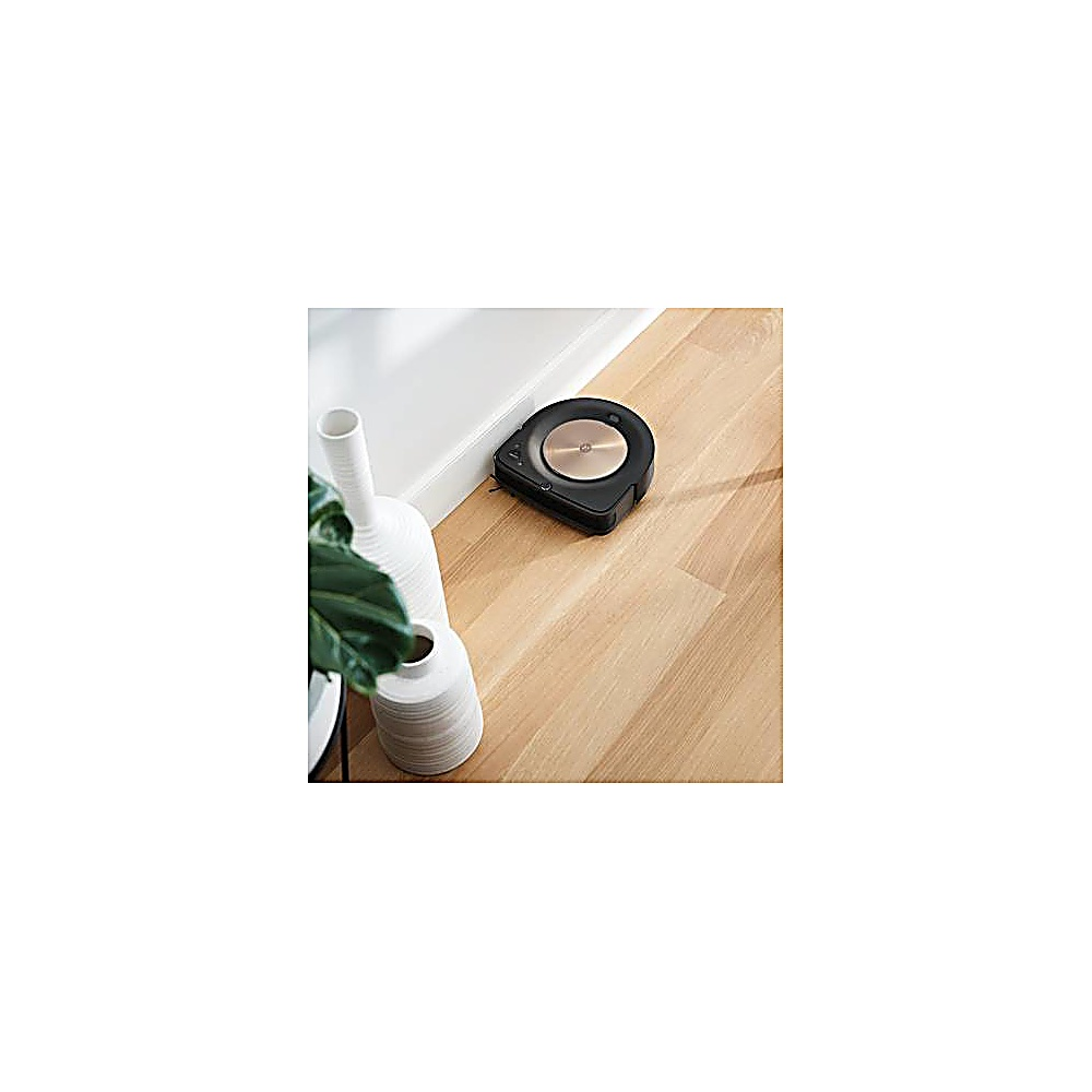 Ein schwarzer Roboter-Staubsauger auf einem Holzboden in der Nähe einer weißen Wand und Vasen mit grünen Blättern.