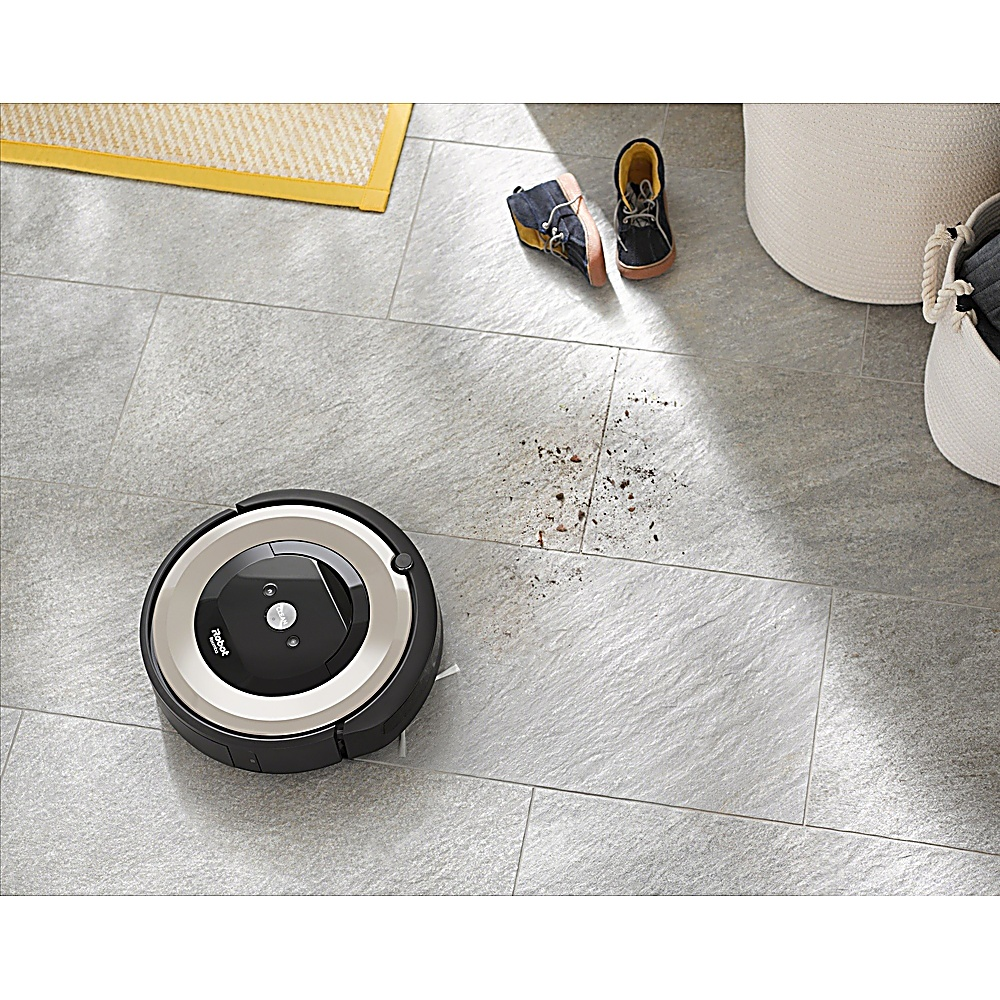 Un robot aspirapolvere su un pavimento piastrellato grigio, vicino a scarpe e cesti. È visibile dello sporco. Un tappeto giallo è sullo sfondo.