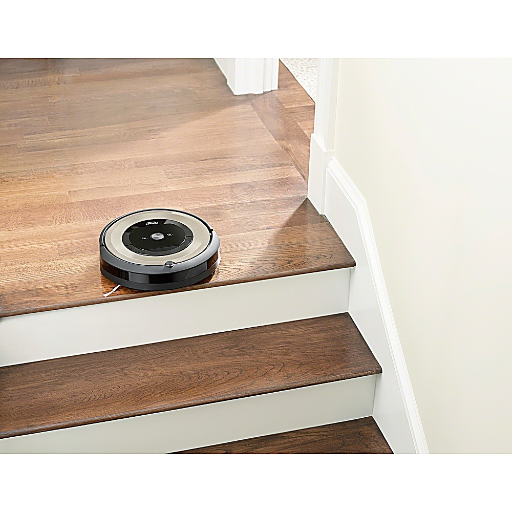 Un robot aspirapolvere su un pavimento in legno vicino alle scale. Le scale hanno alzatine bianche e gradini marroni. Un muro bianco sulla destra.