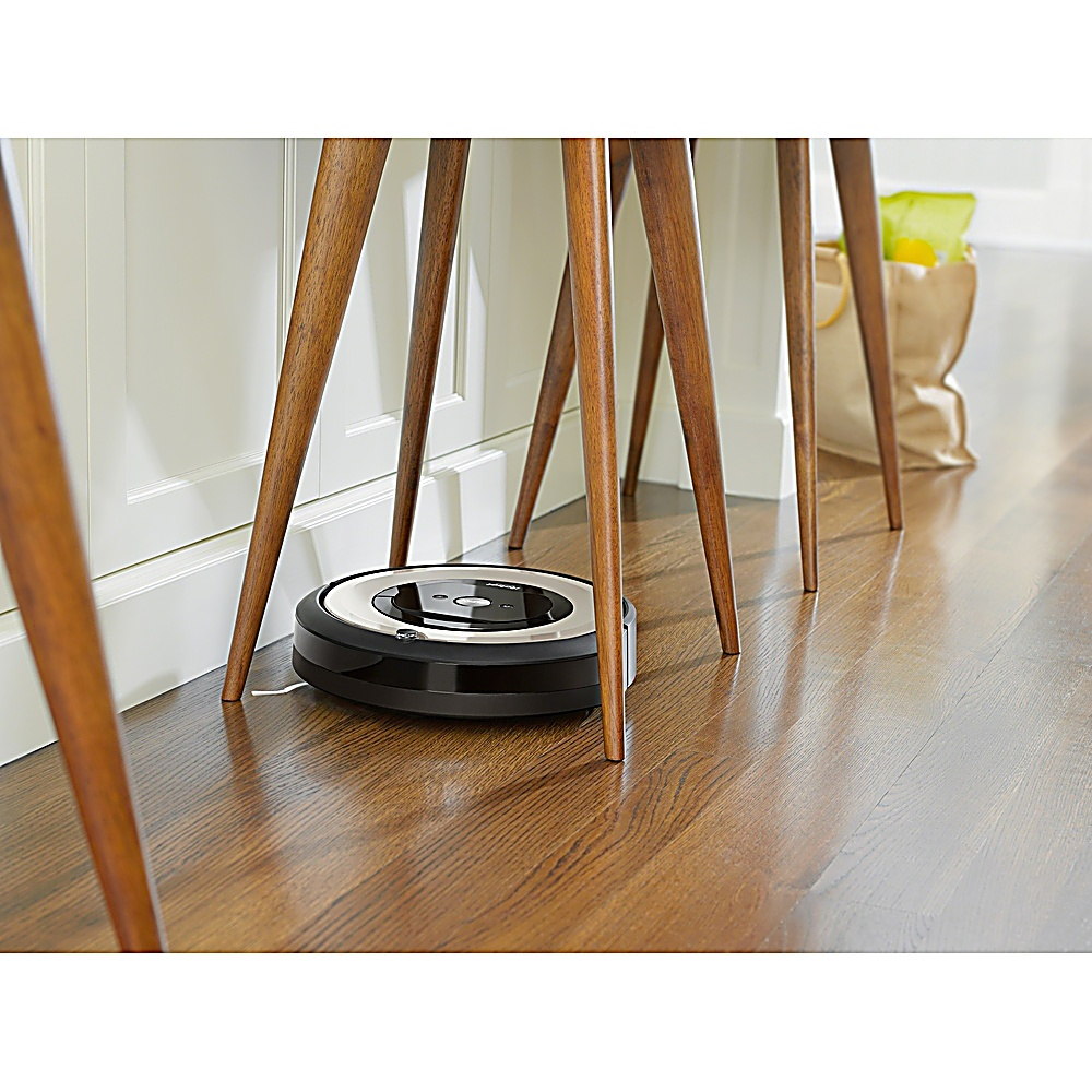 Un aspirapolvere robot sotto un tavolo con gambe in legno. Una borsa della spesa è sullo sfondo.